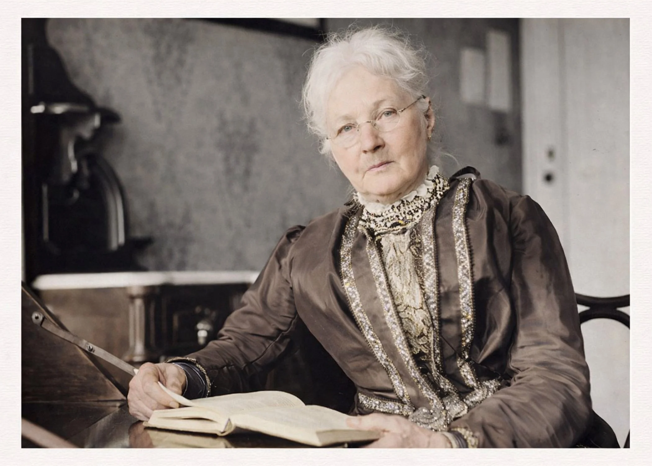 An elderly woman with white hair, wearing glasses and a detailed vintage-style dress, is reading a book at a table in a room with a gray wall and a piano in the background.