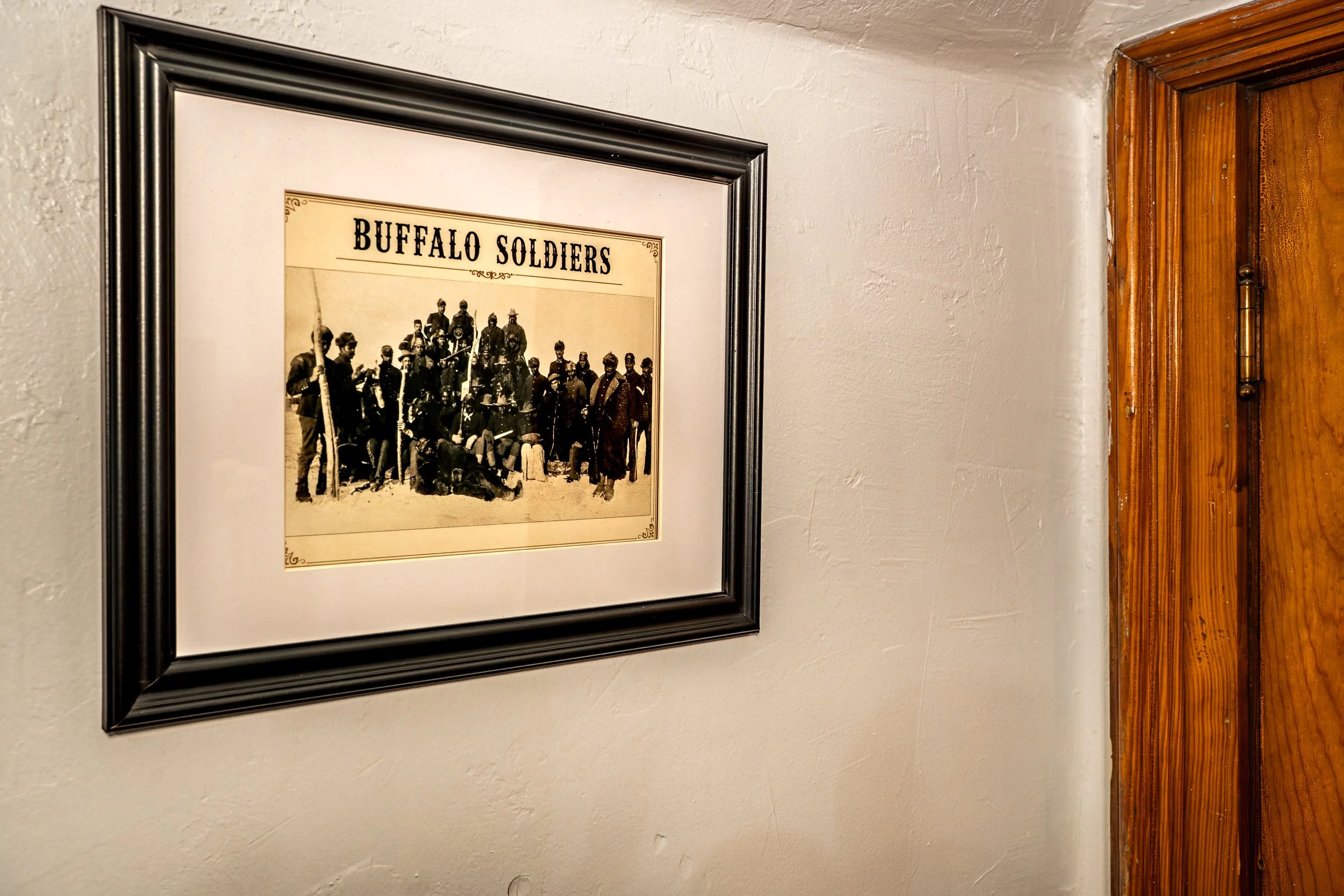 A black frame containing a vintage-style photograph titled 'Buffalo Soldiers' hangs on a white textured wall near a wooden doorframe.