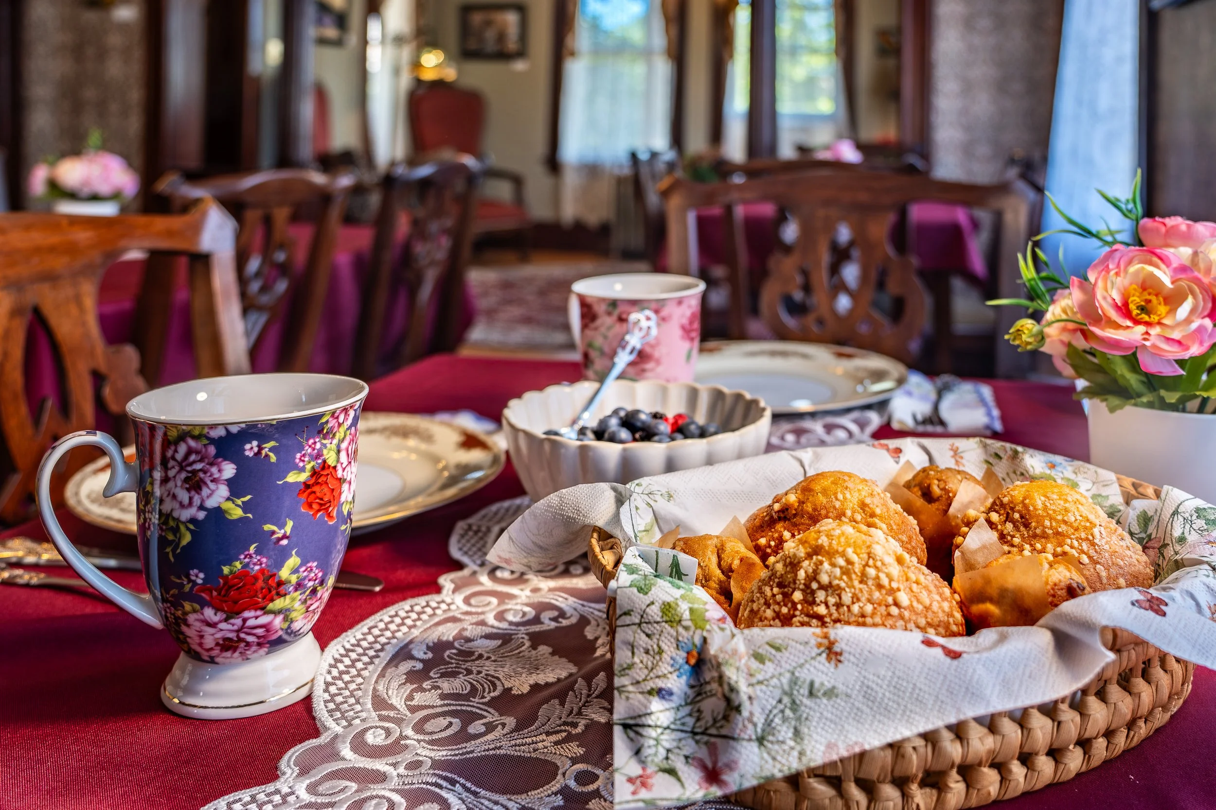 A table set for a meal with floral patterned cups, a bowl of blueberries, a basket of glazed pastries, and a vase of pink flowers, in a cozy dining room with wooden chairs and windows.