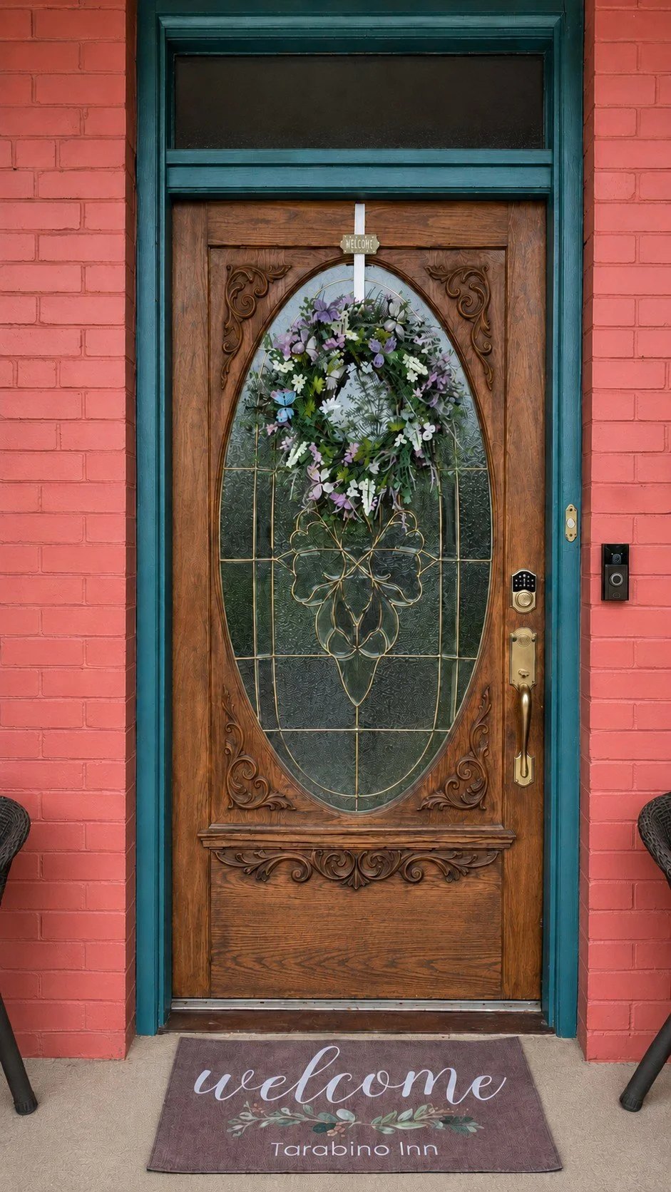 Decorative wooden front door with an oval glass panel, etched floral and vine design, and a floral wreath hanging in the center. The door has ornate carved wood trim and is framed by red brick walls and teal trim. A welcome mat reading "welcome Tarabino Inn" is placed in front.