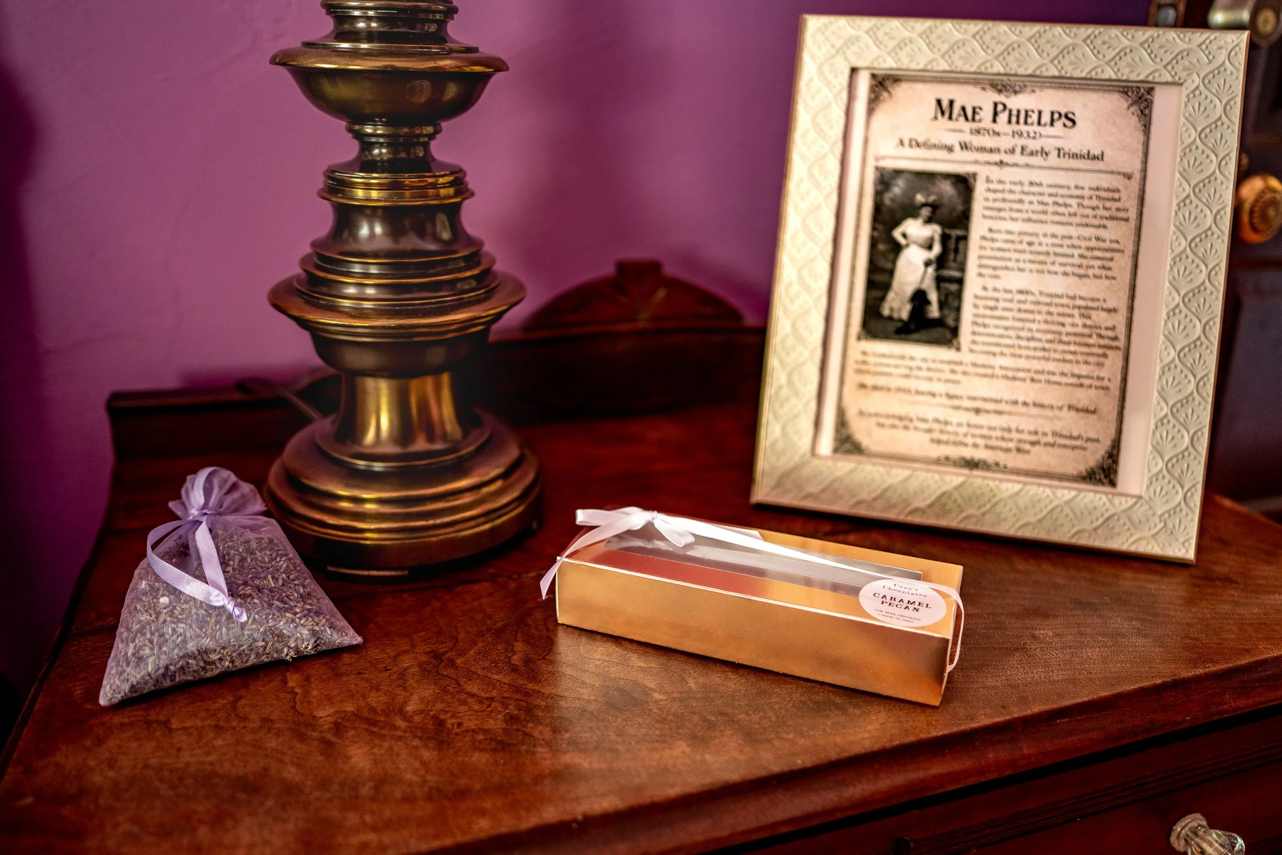 A wooden table displaying a brass candlestick holder, a framed biography of Mae Phelps, a small lavender bag, a box of caramel pagan chocolates, and a small bag of lavender.