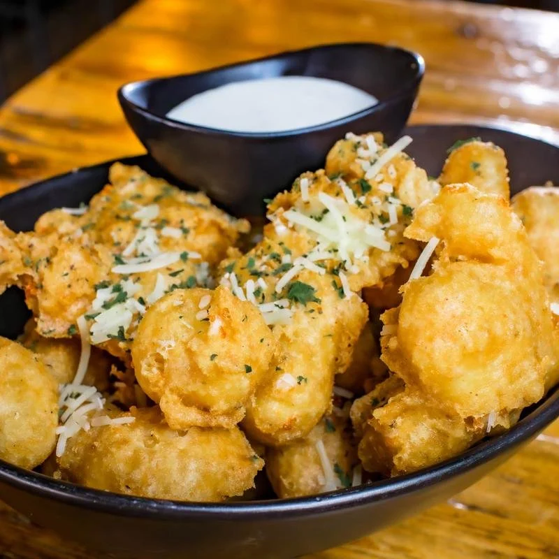 Bowl of fried cauliflower florets topped with shredded cheese and herbs, with a small dish of dipping sauce on a wooden table.