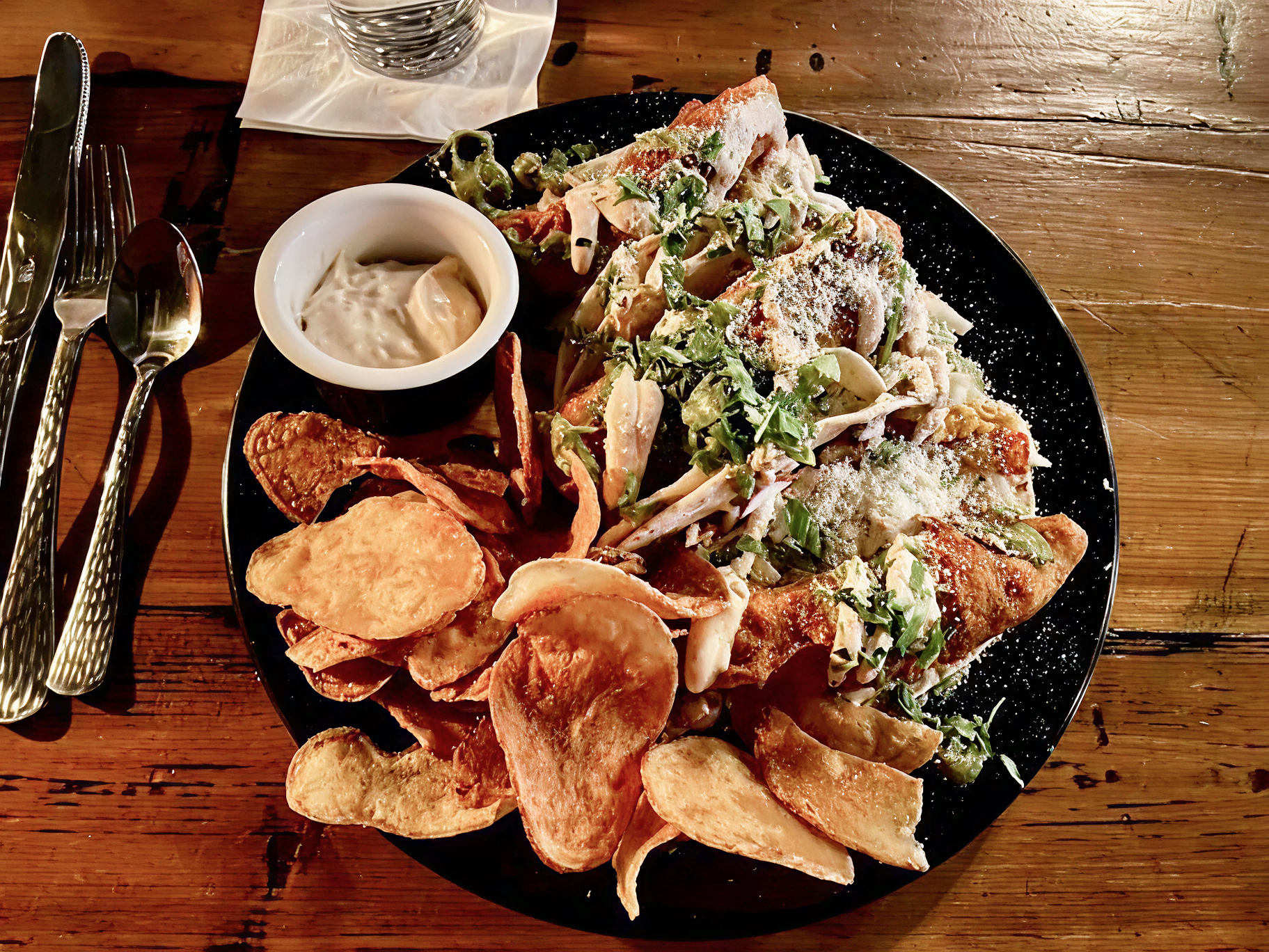 Plate of nachos with chips, melted cheese, guacamole, sour cream, and chopped greens, served on a black plate on a wooden table.