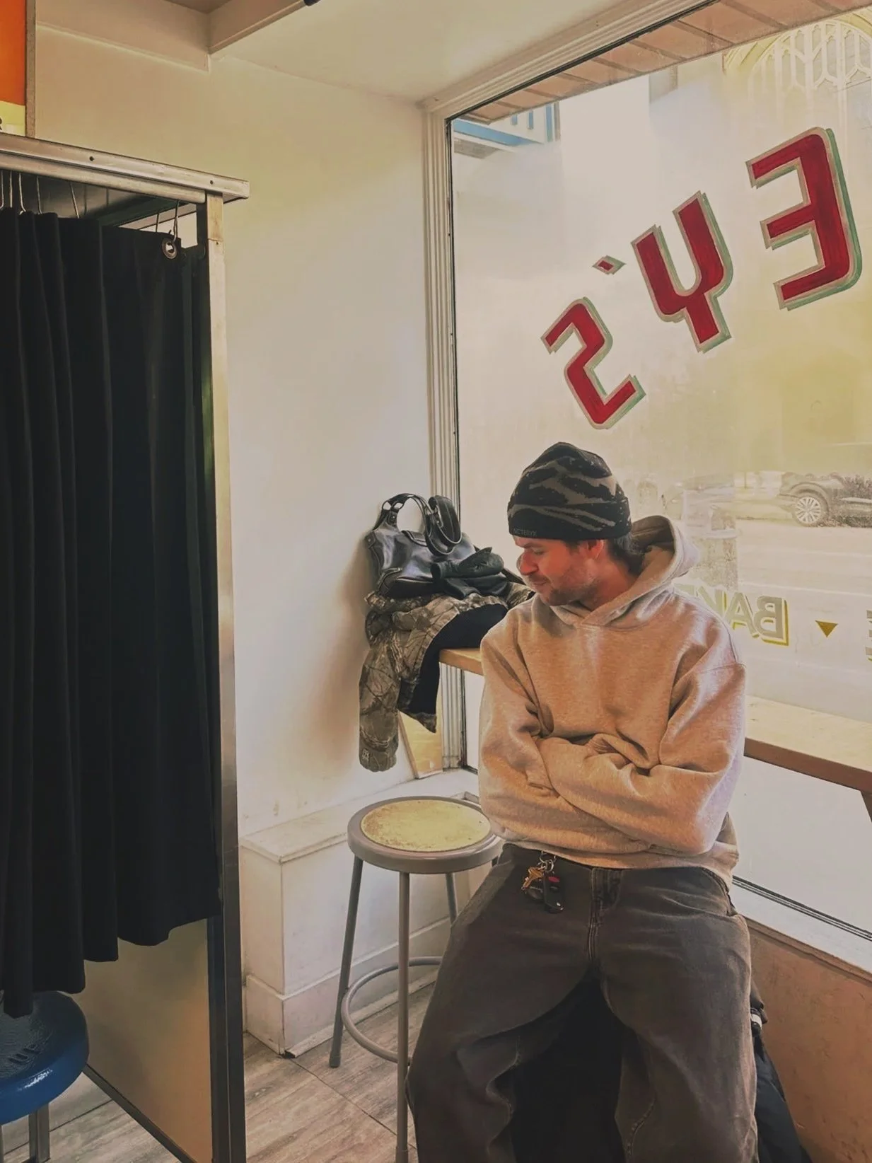 A man sitting with arms crossed inside a cafe with large windows, wearing a gray hoodie and beanie, next to a counter with bags and a jacket hanging on a hook, with a sign in reverse on the window reading 'CUE 24'.