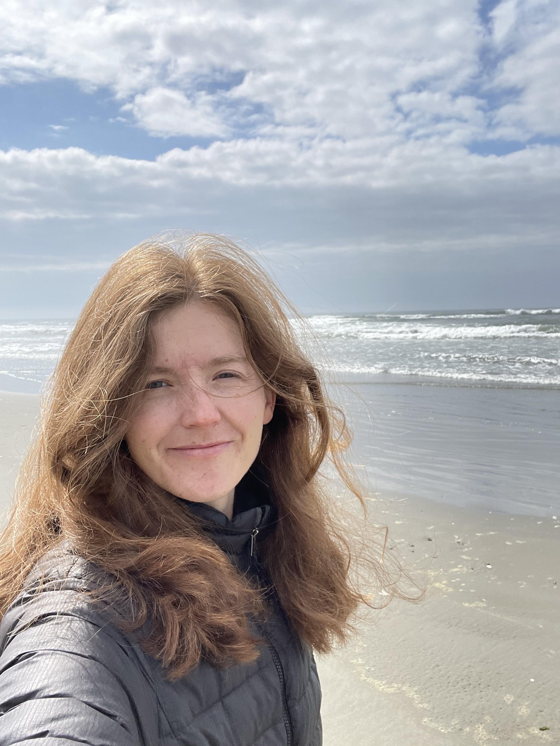 A woman with long red hair smiling at the camera while standing on a sandy beach with waves and a blue sky with clouds in the background.
