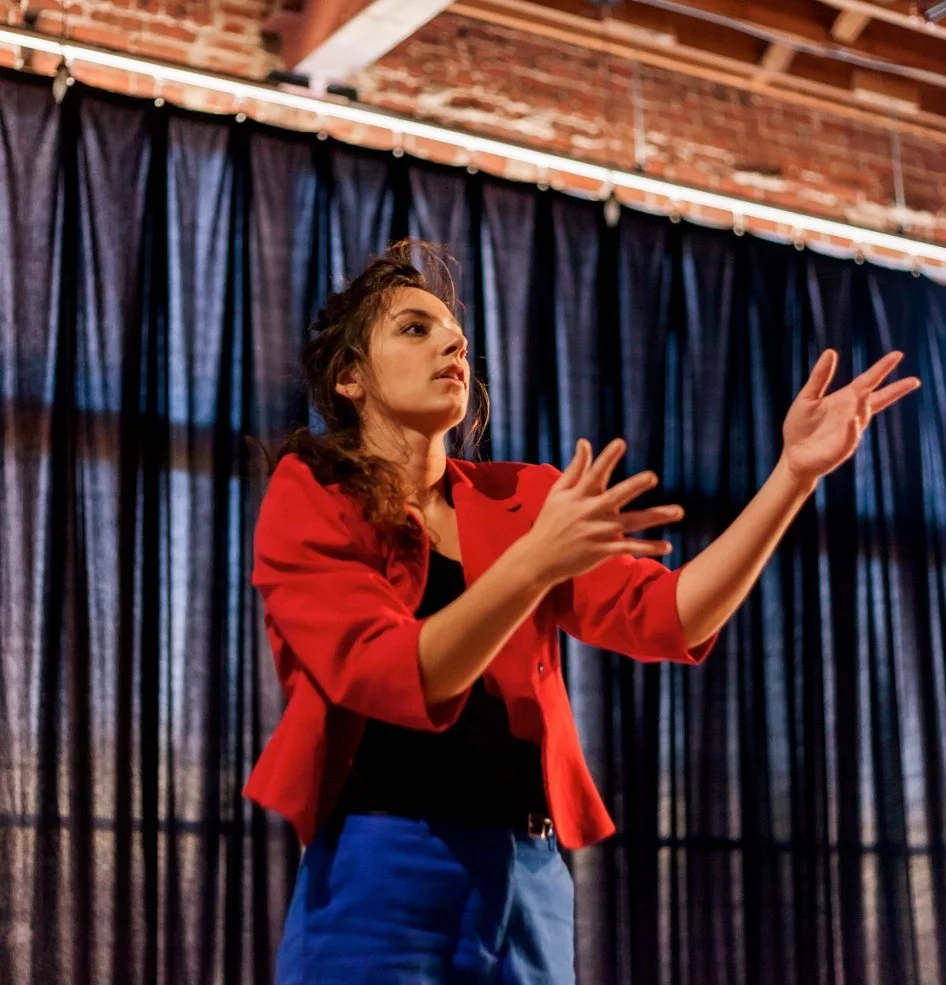 Anna gestures upwards with her arms. She wears red and blue.