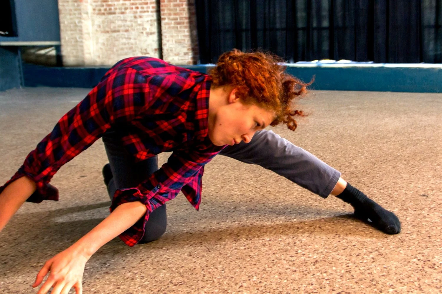 Leah dances on the floor at Motorco. They wear  a red and black plaid shirt, grey pants, and black socks.