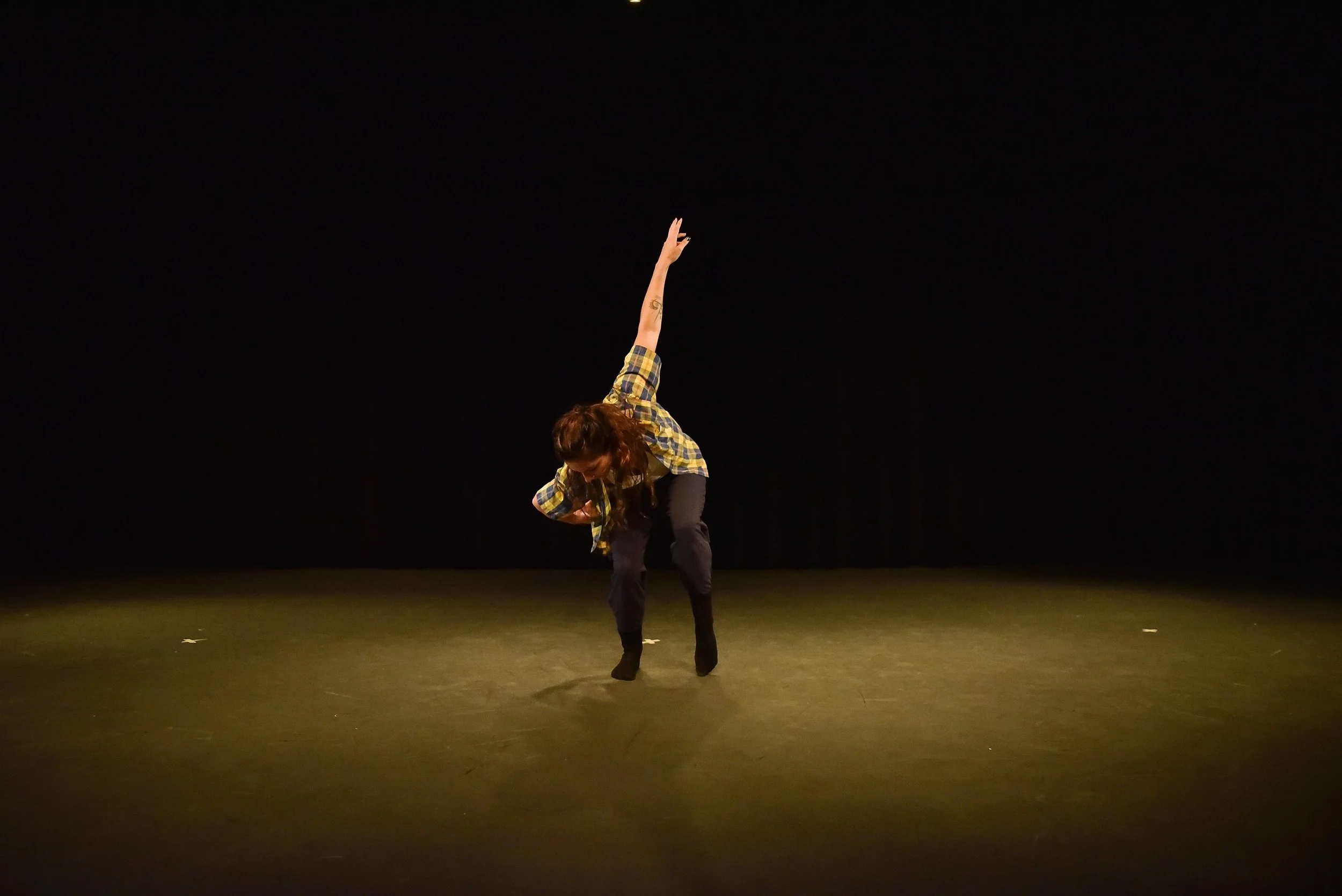 Anna leans forward with one arm down and one arm reaching up on an angle. She wears a yellow and blue plaid short sleeved shirt over a white tee shirt and black pants. 