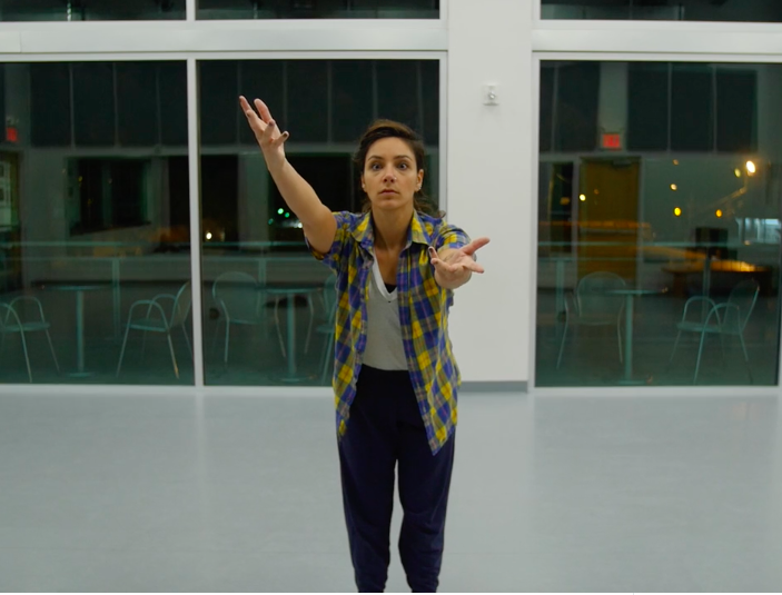 Anna leans forward with arms outstretched on a high angle. She wears a yellow and blue plaid short sleeved shirt over a white tee shirt and black pants. She is in a dance studio.