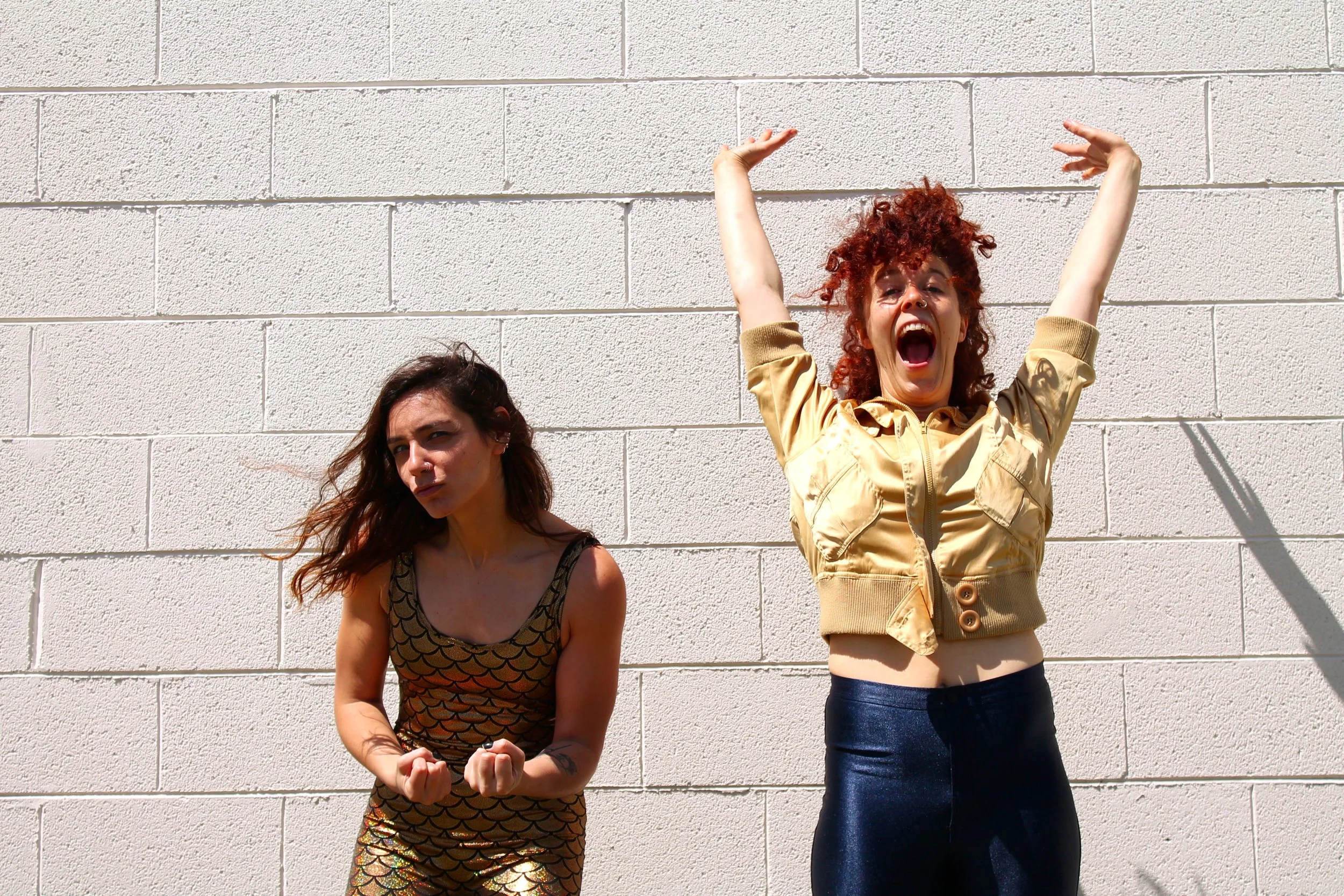 Anna and Leah pose in front of a white brick wall. Anna wears a gold unitard with a black fish scale design. Leah wears a yellow gold jacket and shiny blue pants. She reaches upwards. 