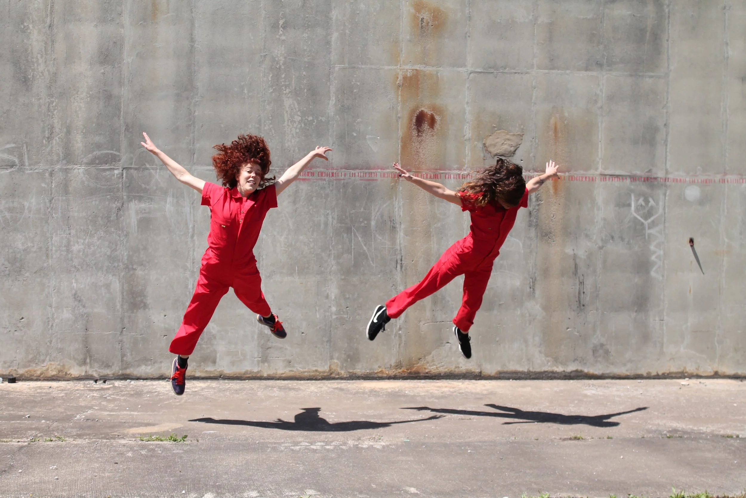 Leah and Anna wear red jumpsuits. They are doing flying squirrel jumps in front of a grey concrete wall.