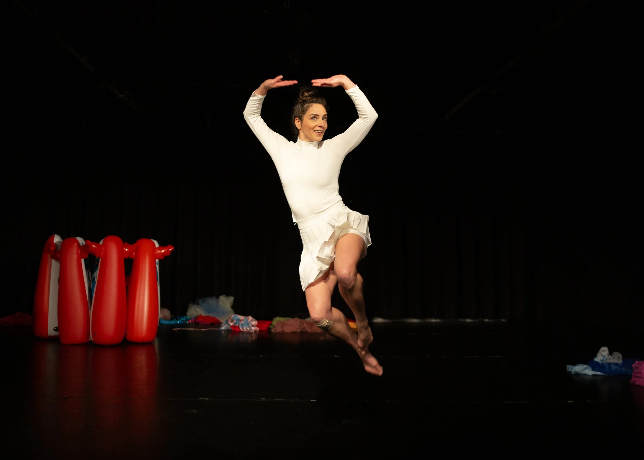 Anna wears all white. She leaps with bent legs and arms overhead. Inflatable Bozo dolls are turned away from her upstage.