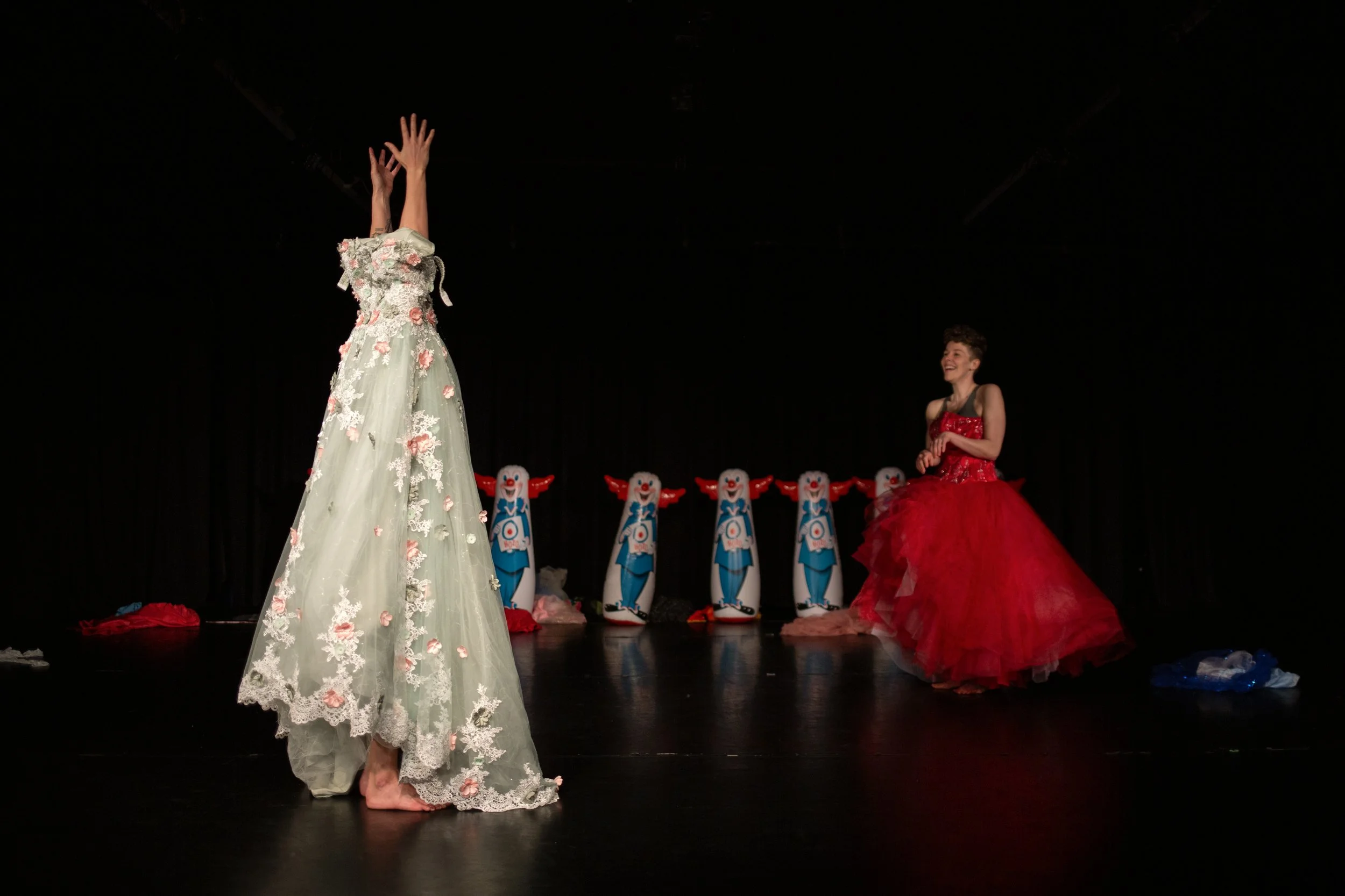 Anna stands with arms overhead, putting on a long formal dress. Leah stands behind her with a red formal poofy dress on. Inflatable Bozo dolls line the back of the stage. 