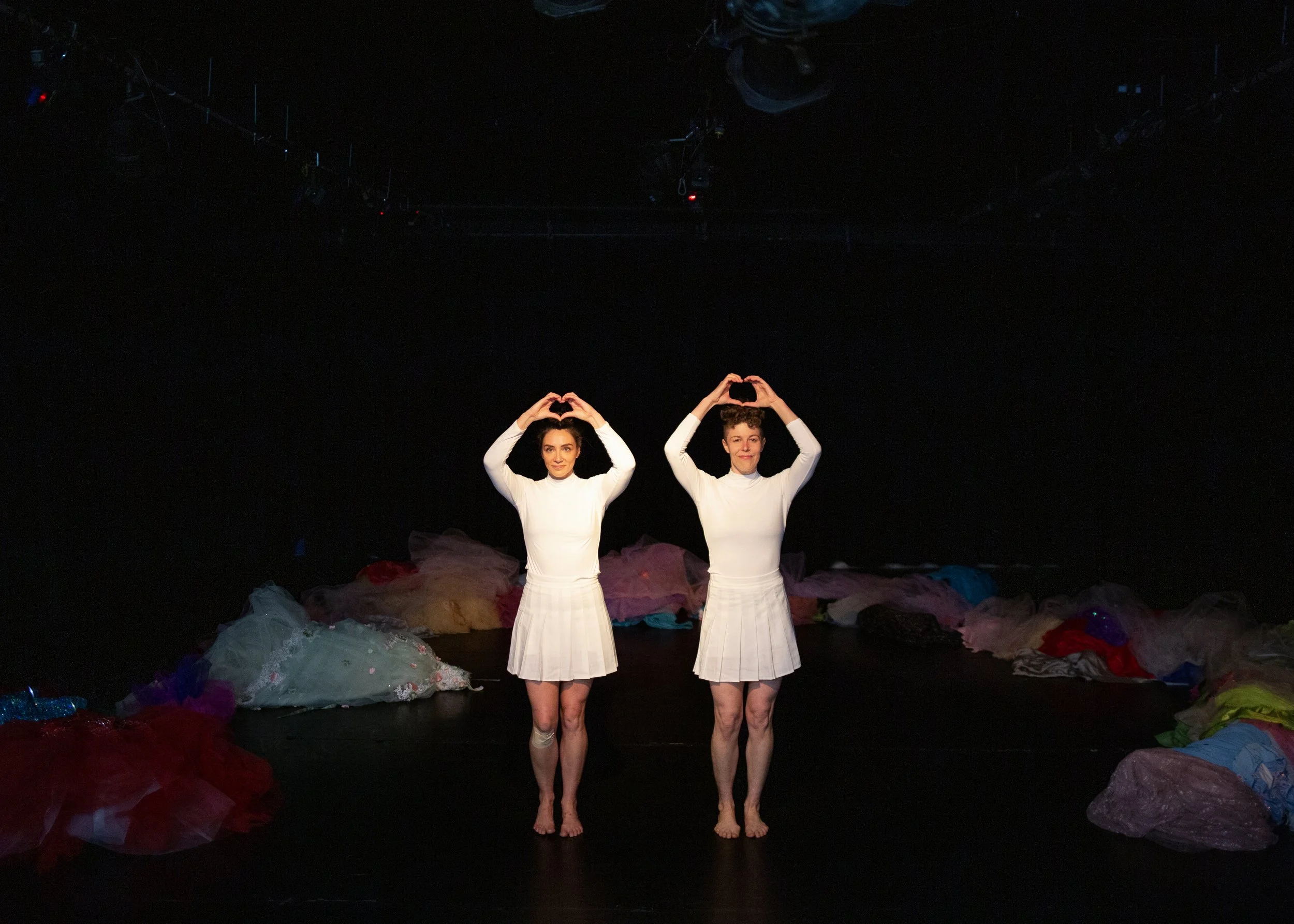 Anna and Leah wear all white outfits. They make heart gestures with their hands overhead. The stage is lined with colorful tulle.