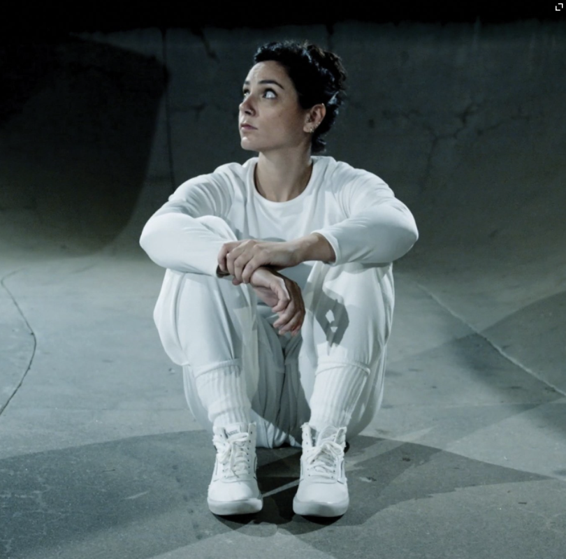 Anna wears all white. She pulls her knees into her chest, staring up toward the corner. She is in a skate park bowl.