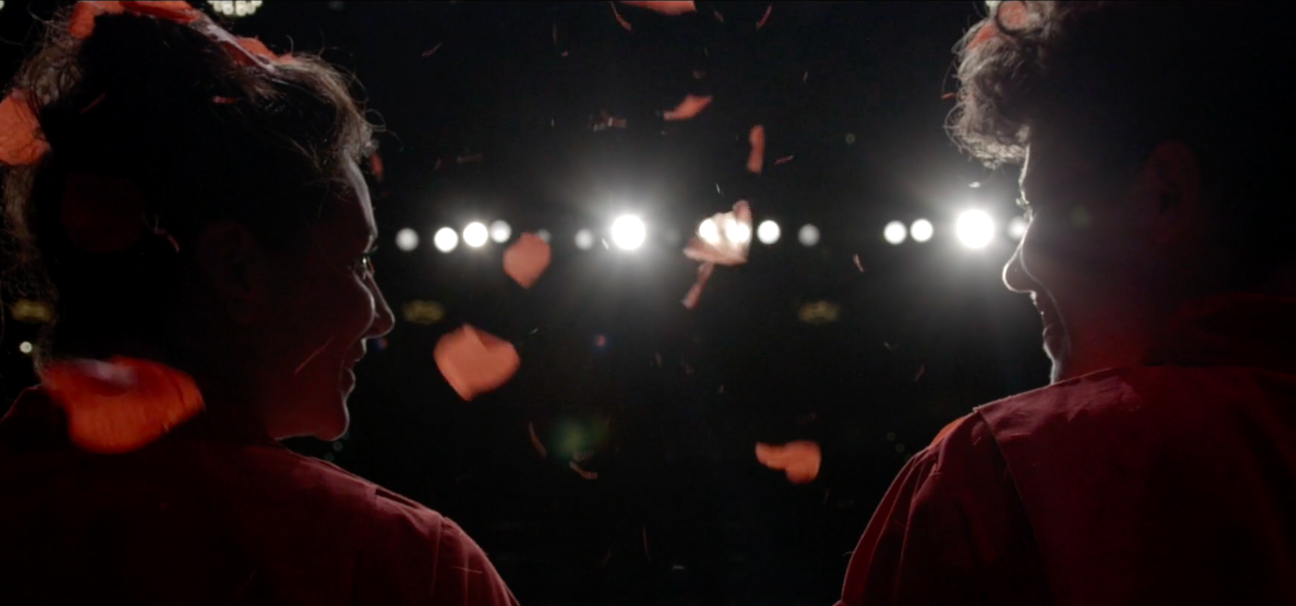 Anna and Leah are onstage and can be seen from behind, rose petals falling down on them as they look at one another.