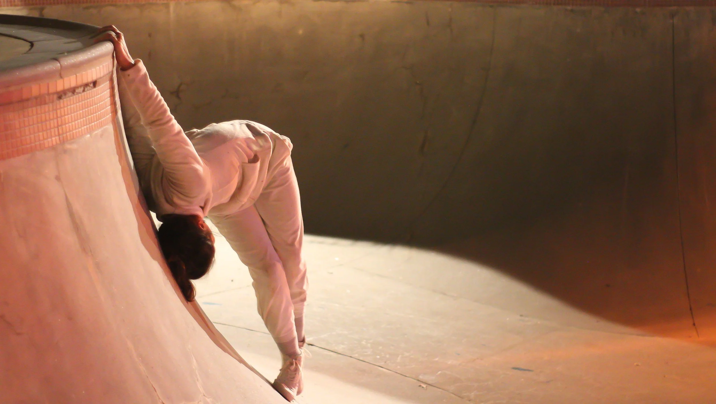 Anna dances in a skate park bowl. She wears all white and holds onto the ledge.
