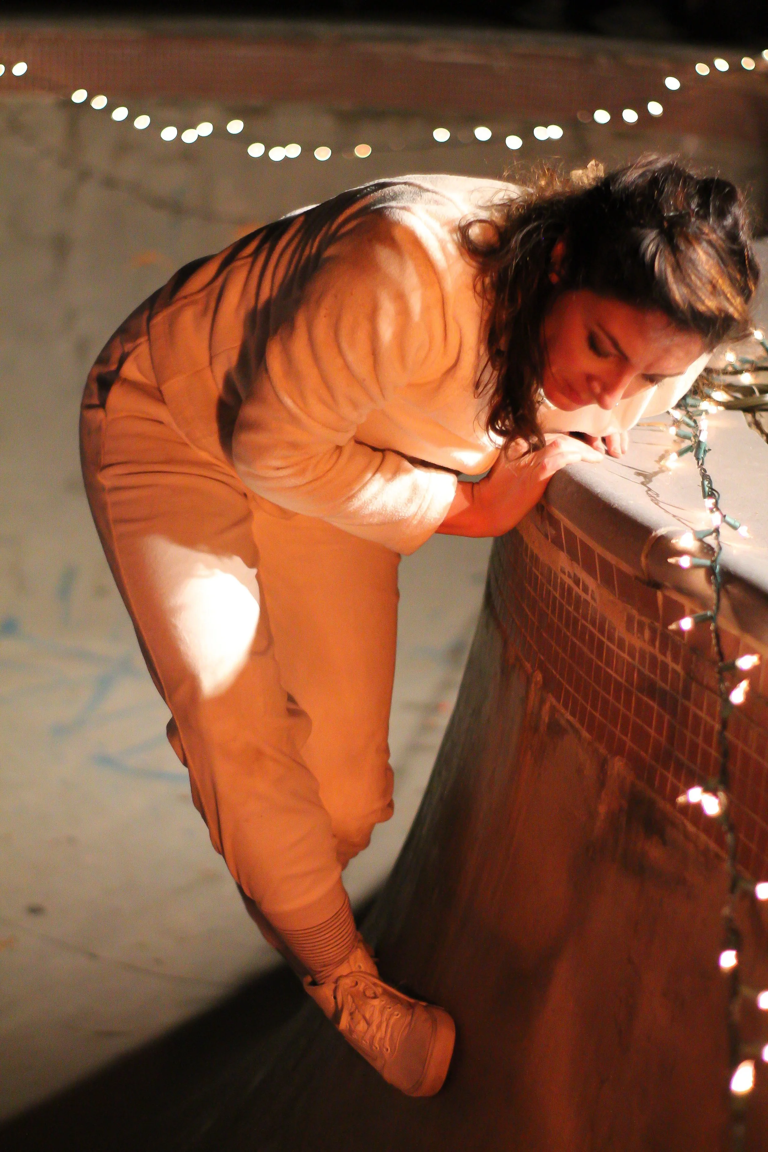 Anna wears all white. She clings to the ledge of the bowl at the skate park. Twinkle lights are hung along the edge.