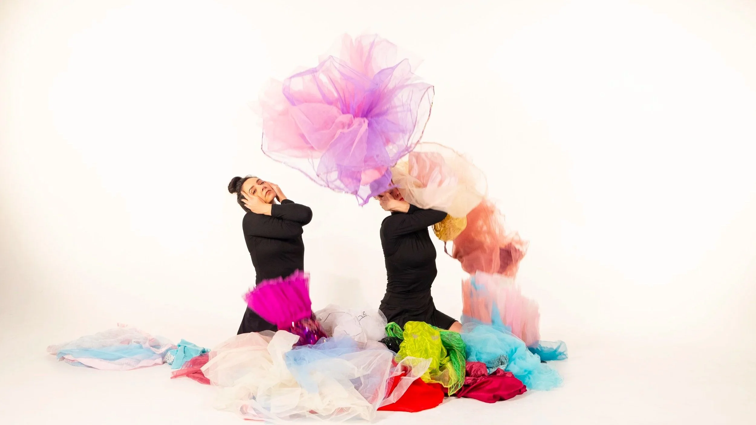 Anna and Leah wear all black. They sit on their shins, and their hands manipulate their faces to look strained. They are surrounded by colorful tutus, some of which are lifting off the floor.