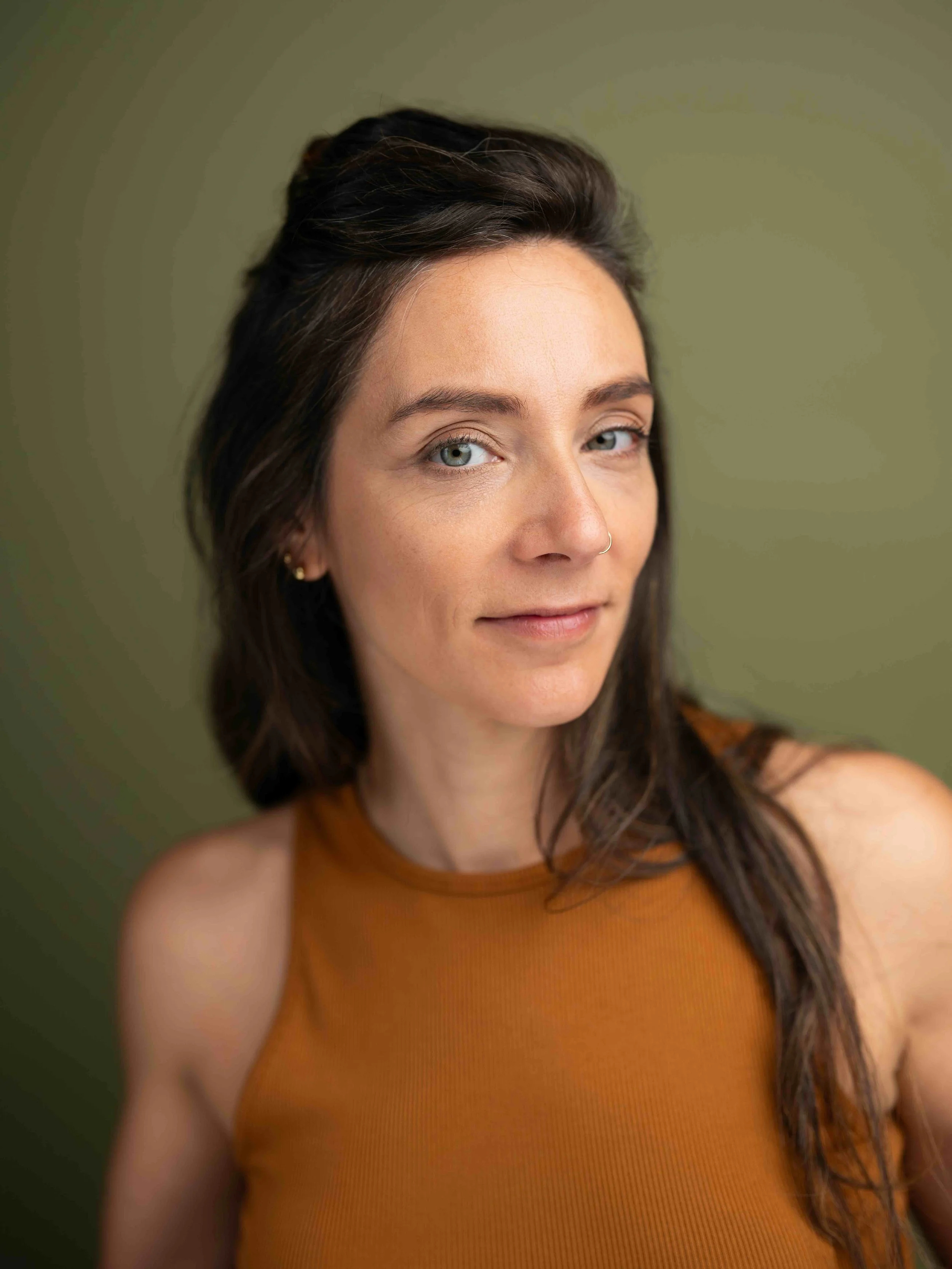 Anna wears an orange tank top. She looks at the camera while standing in front of a green background.