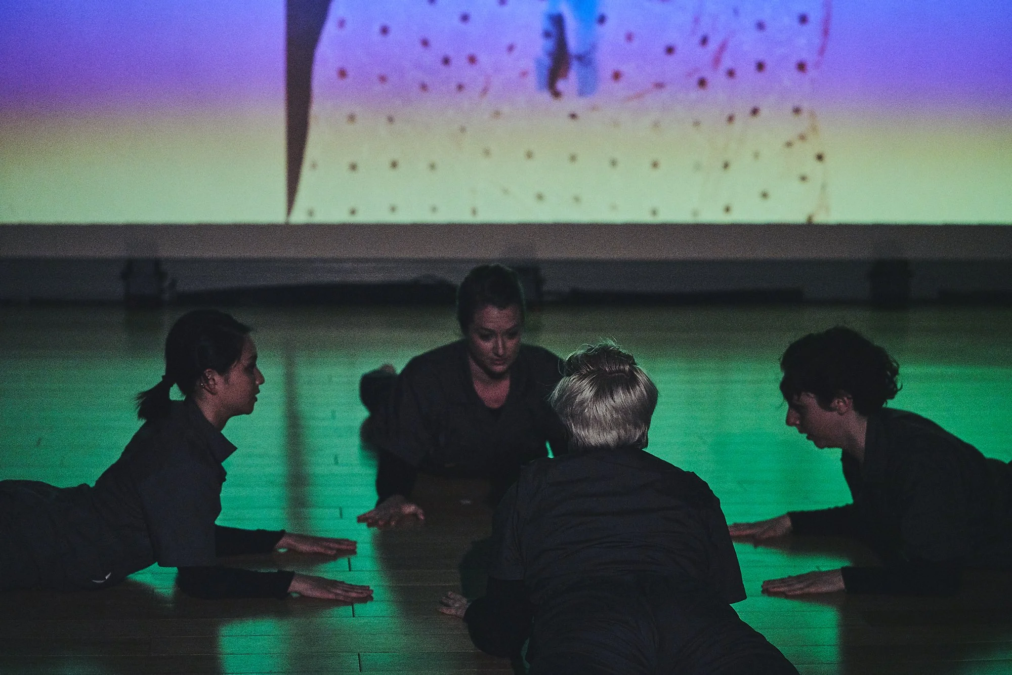 Four dancers lie on their stomachs, facing one another. A multicolored lighting scheme can be seen behind them. 