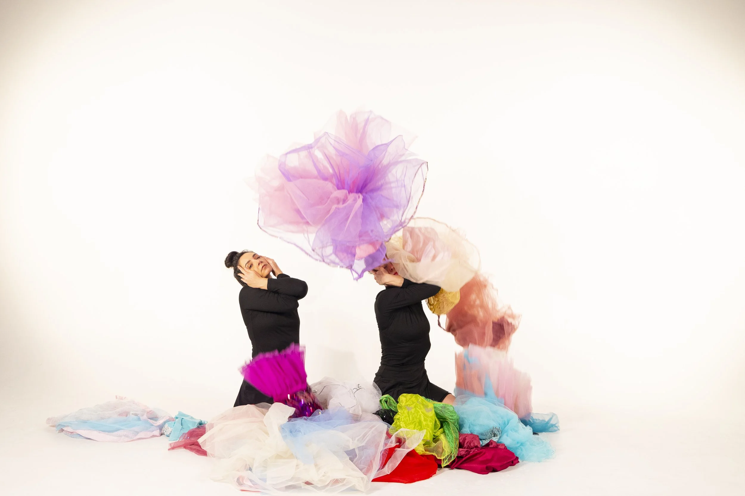 Anna and Leah wear all black. They sit on their shins, and their hands manipulate their faces to look strained. They are surrounded by colorful tutus, some of which are lifting off the floor. 