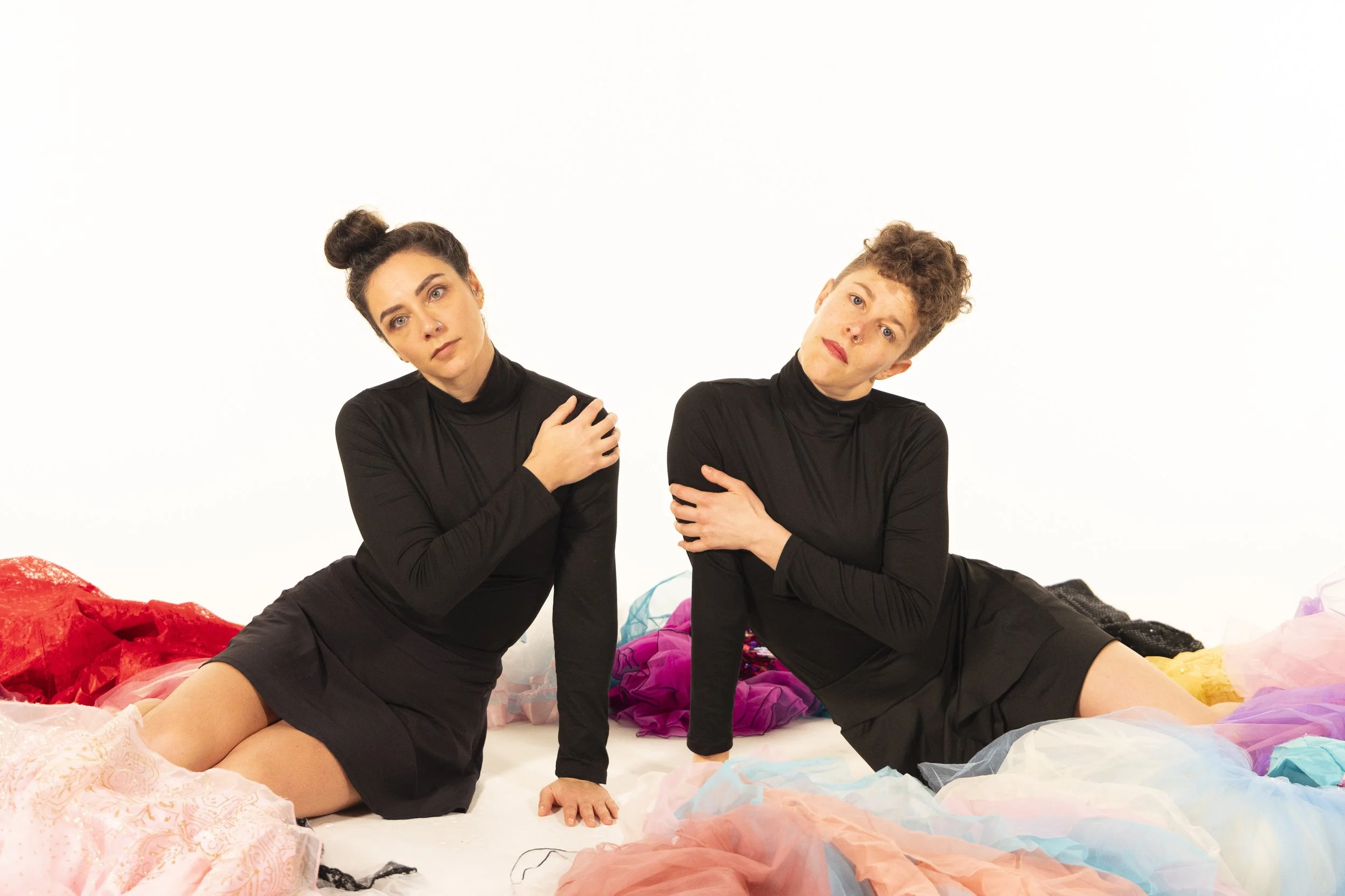 Anna and Leah wear all black. They lean on one arm while seated on the floor, faces expressionless and staring ahead. They are surrounded by colorful tutus. 