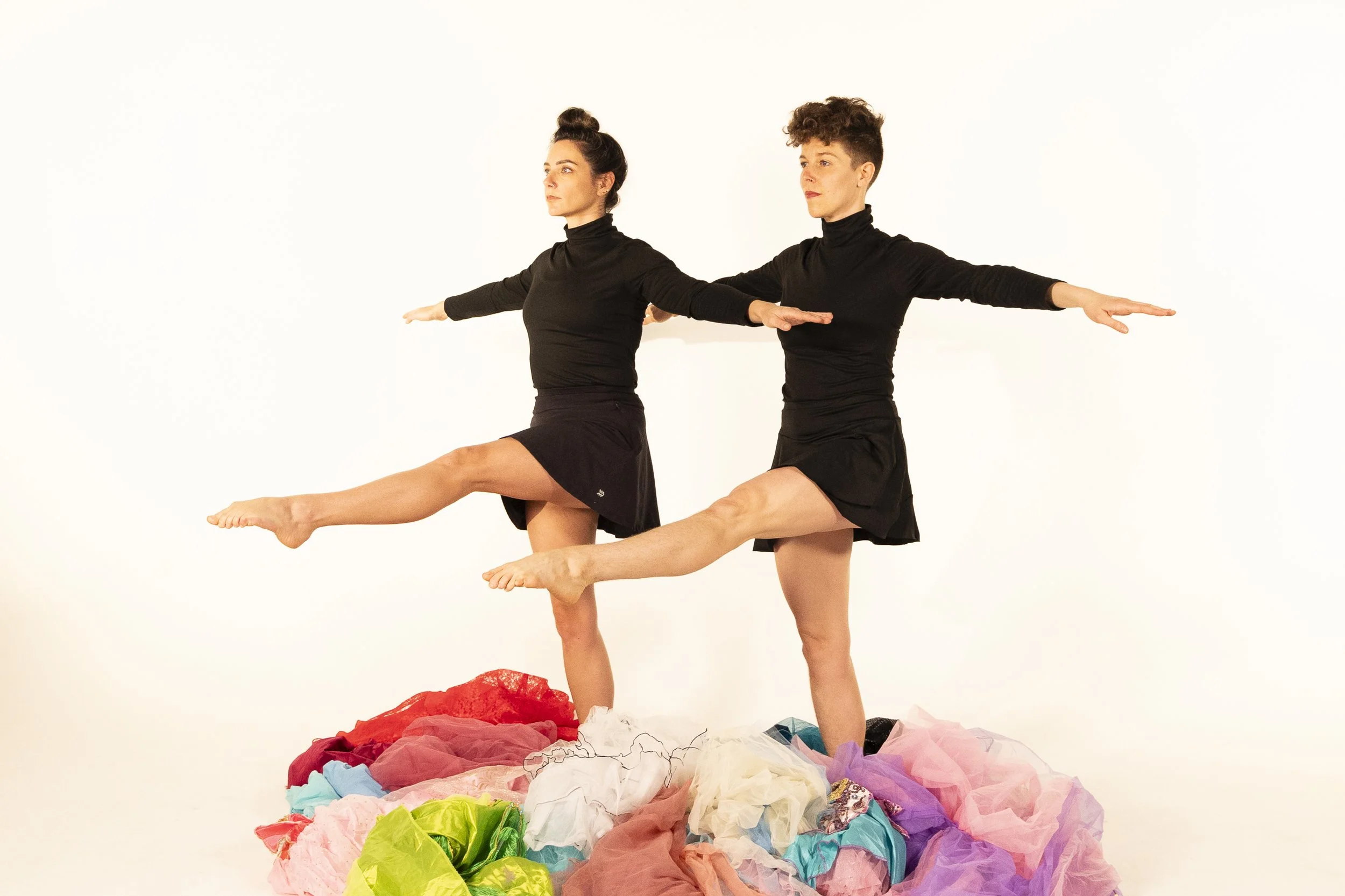 Anna and Leah wear all black. They balance on one leg with arms outstretched, angled toward the corner. They are surrounded by colorful tutus. 