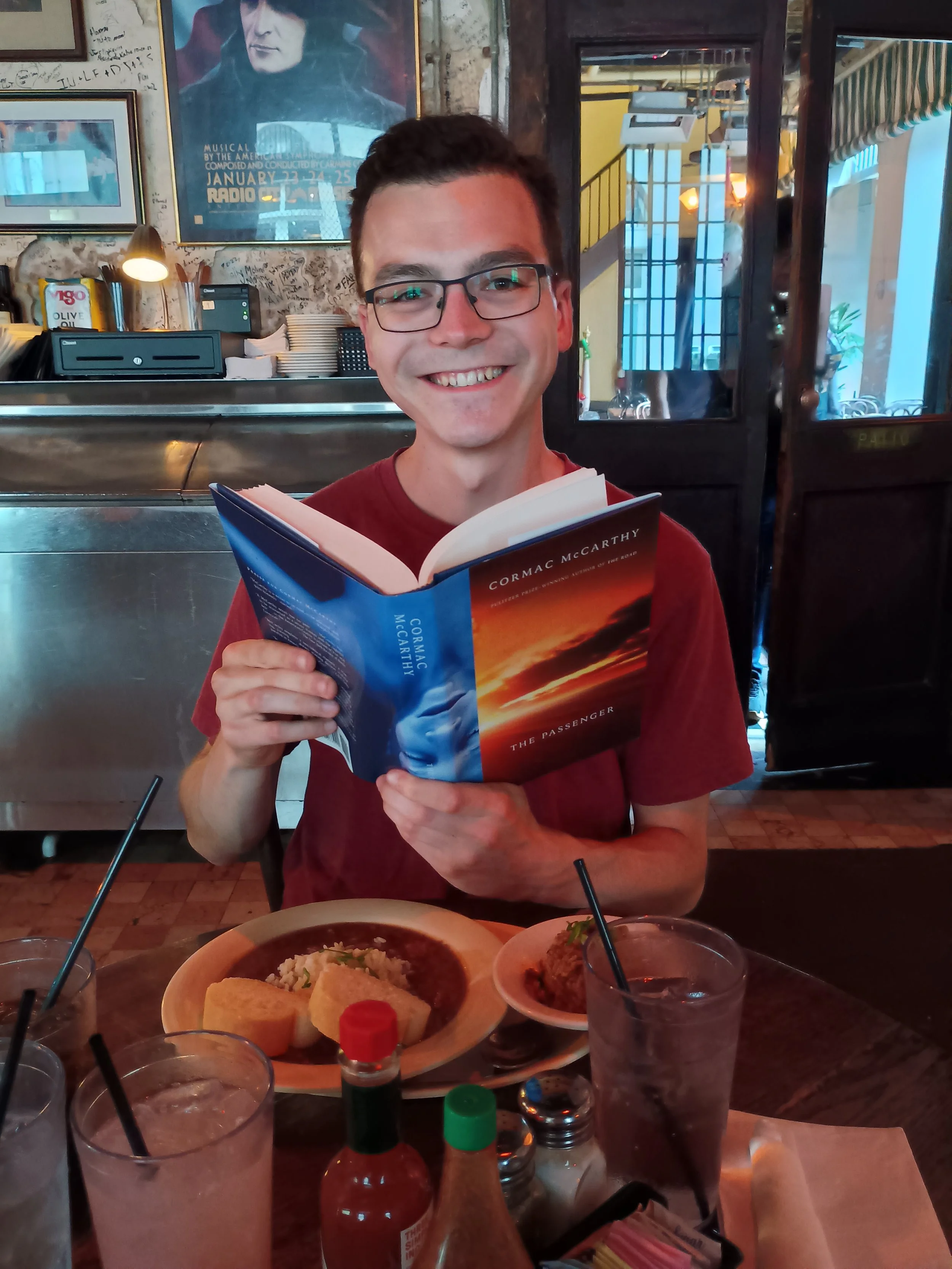 A smiling young man with glasses holding an open book in a restaurant. There are two plates of food, condiments, and drinks on the table in front of him. The background features wall art, including a large framed poster, and a mixed interior decor.