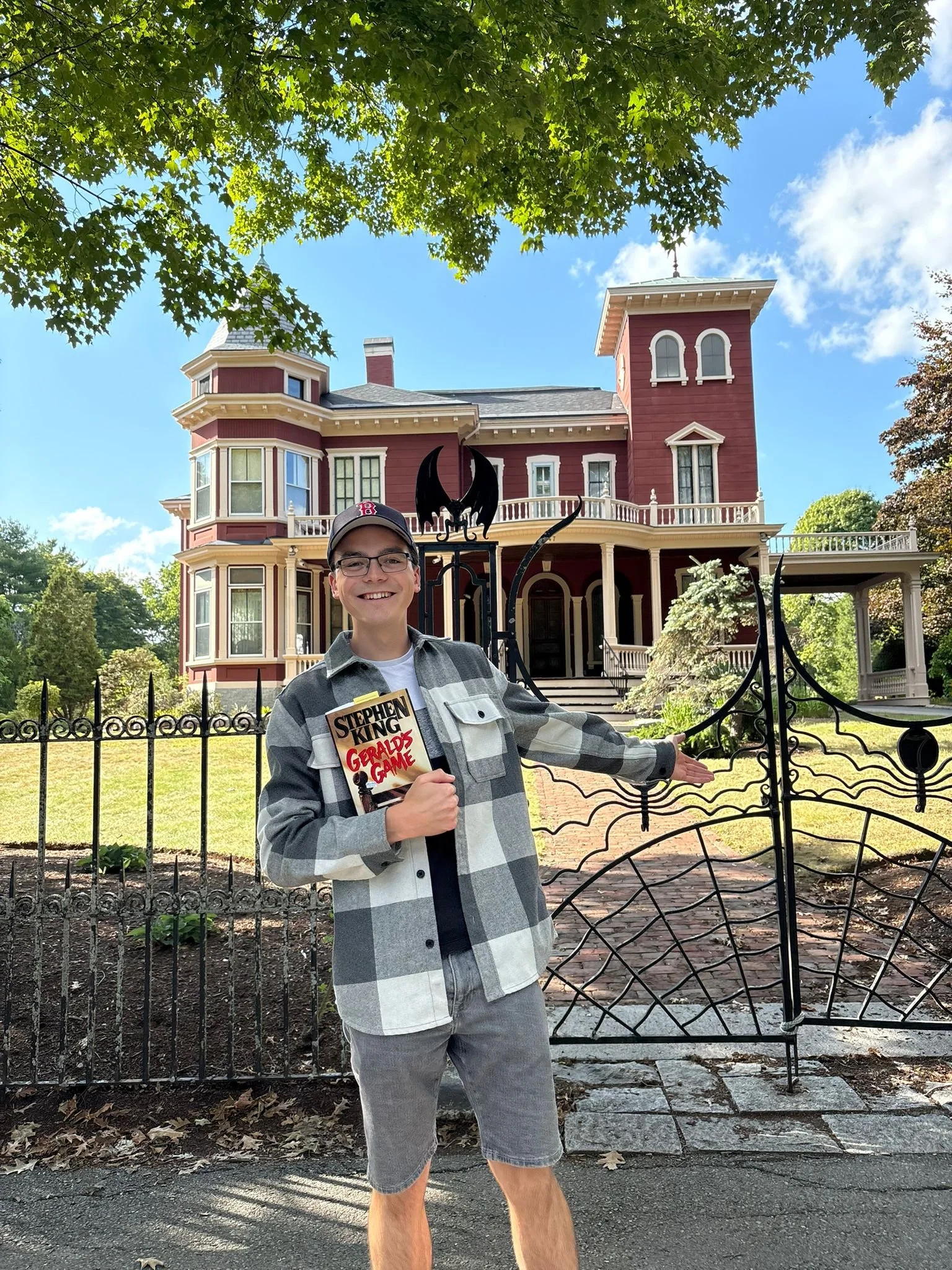 A young man with glasses, wearing a baseball cap and a gray jacket, holding a Stephen King book, stands in front of a wrought iron gate in front of a large, red Victorian-style house with trees and a blue sky in the background.
