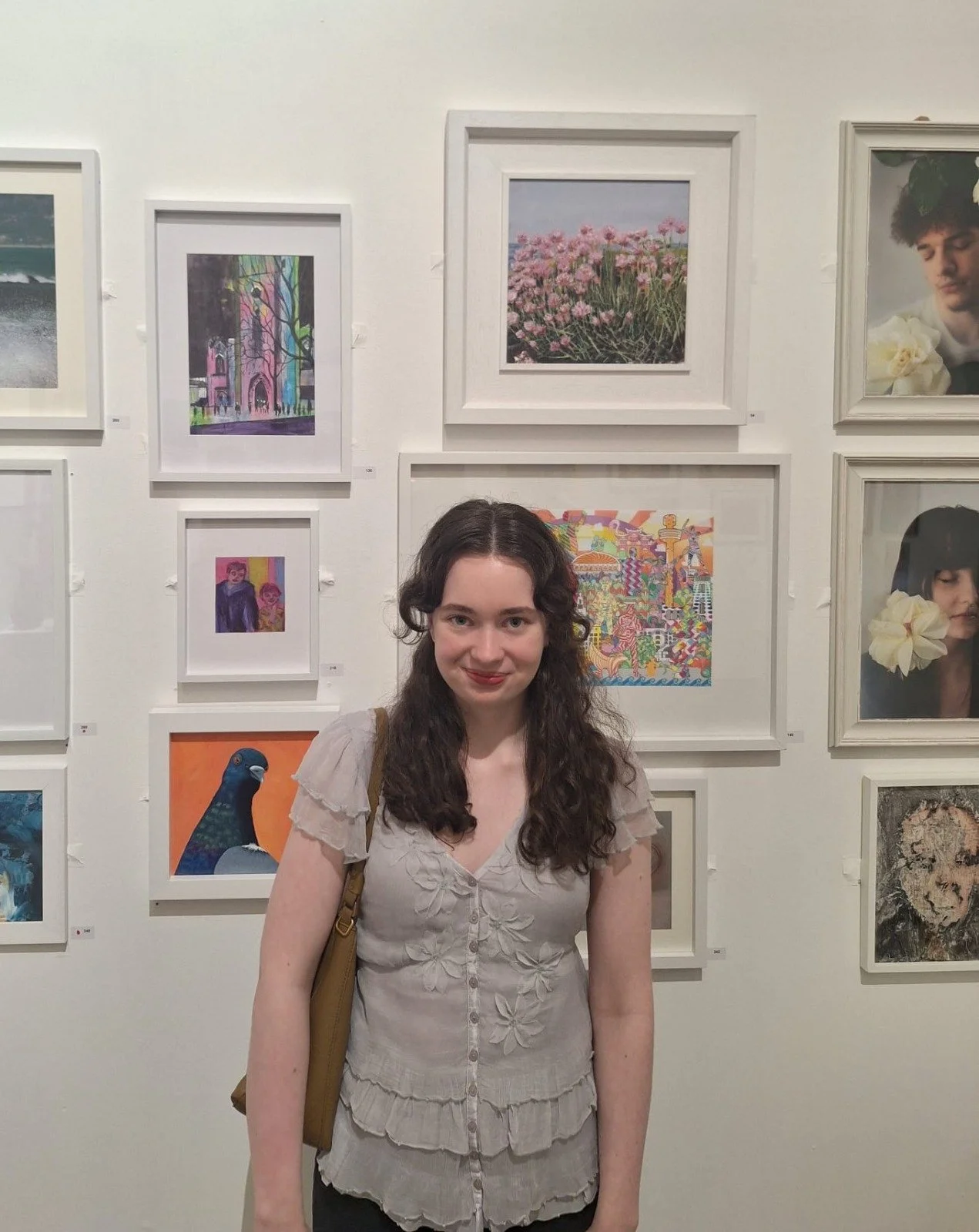 A young woman with long, wavy dark hair, wearing a beige blouse with floral embroidery, standing in front of a white wall with framed artwork at an art gallery.