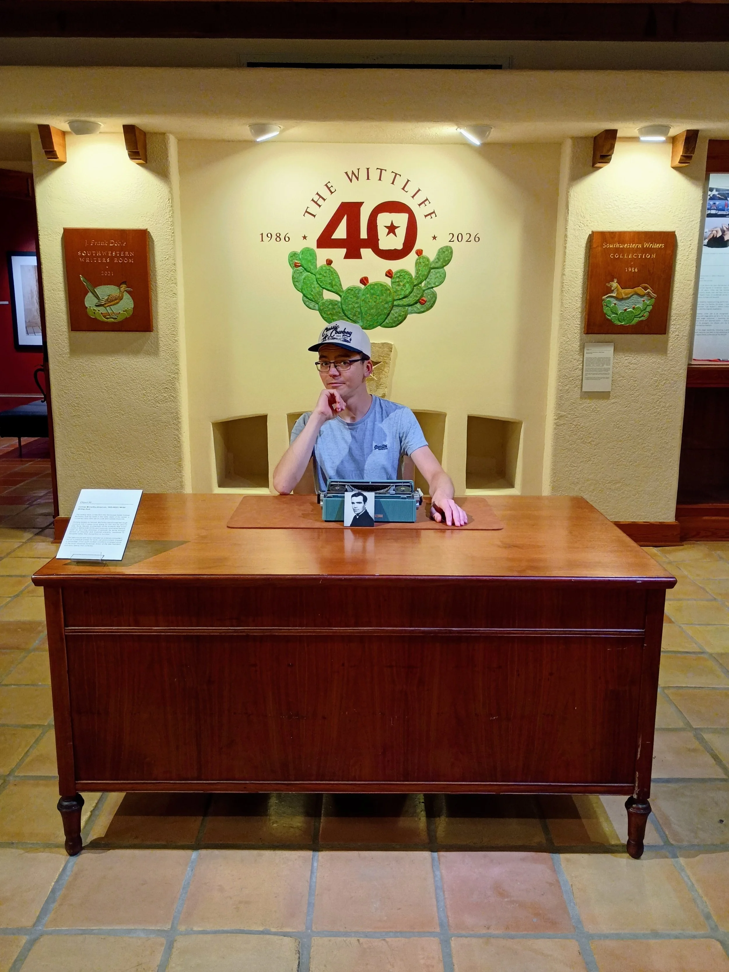 A young man sitting at a wooden desk with a vintage radio and a black-and-white photograph of a man. Behind him, a wall displays a large sign celebrating the 40th anniversary of The Wittlee from 1986 to 2026, with a cactus illustration. Two framed artworks depicting a bird and a horse are on either side of the sign.