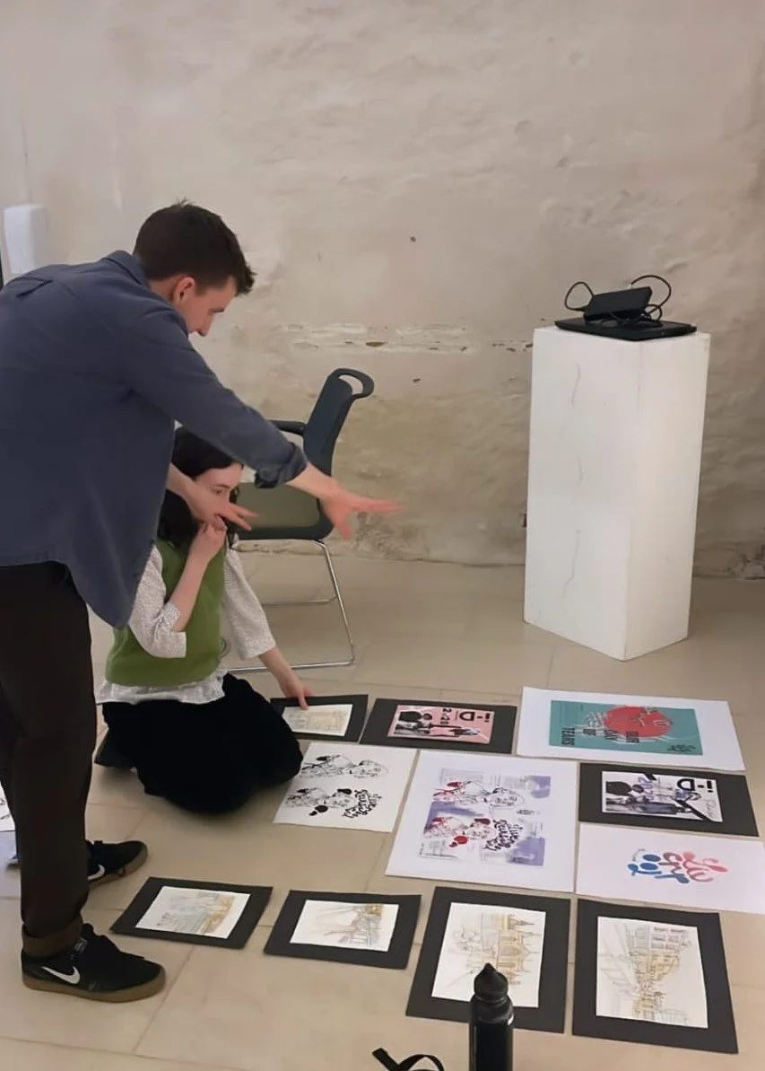 Two people are looking at artwork and photographs laid out on the floor in an art gallery or exhibition space.