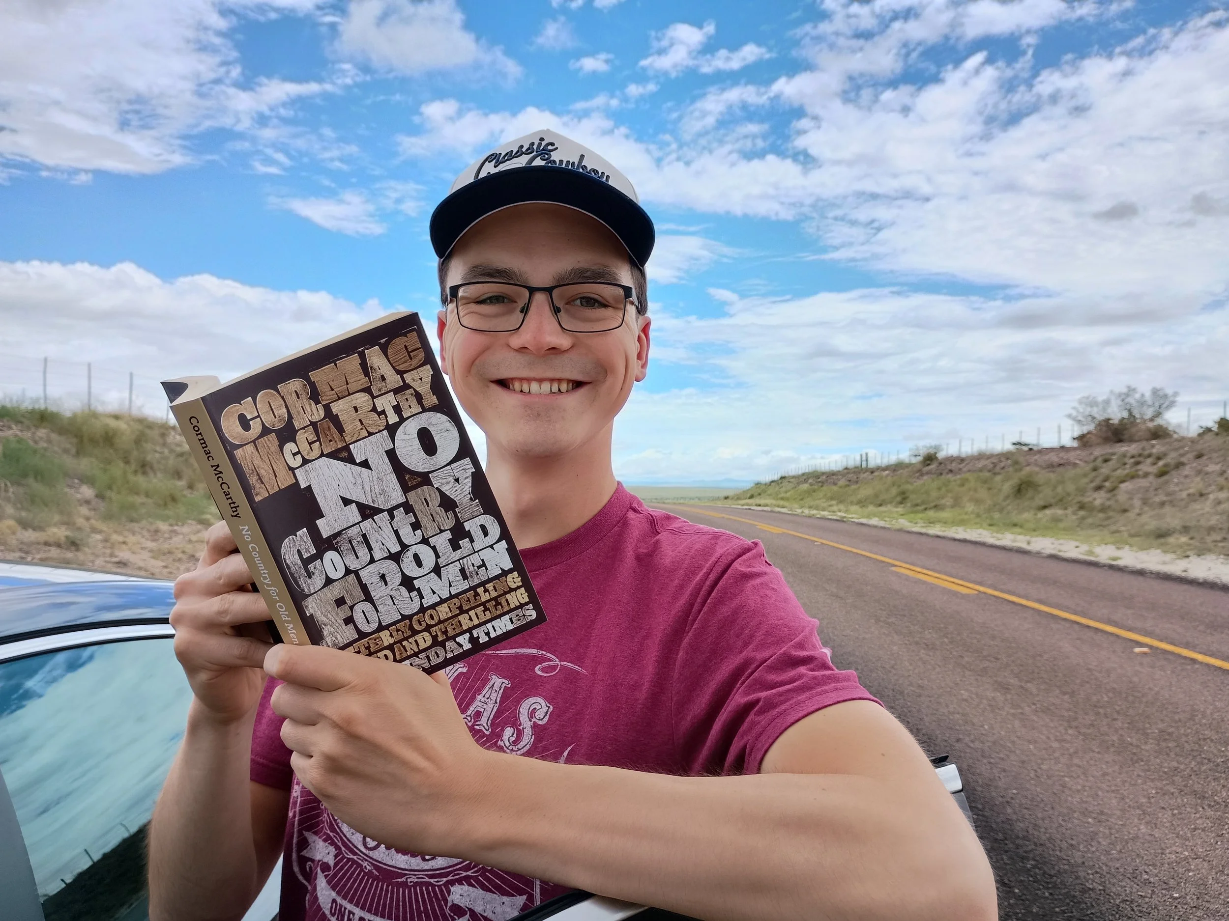 A young man with glasses and a baseball cap smiling at the camera while holding a book called 'Corkboard County' outdoors on a cloudy day.