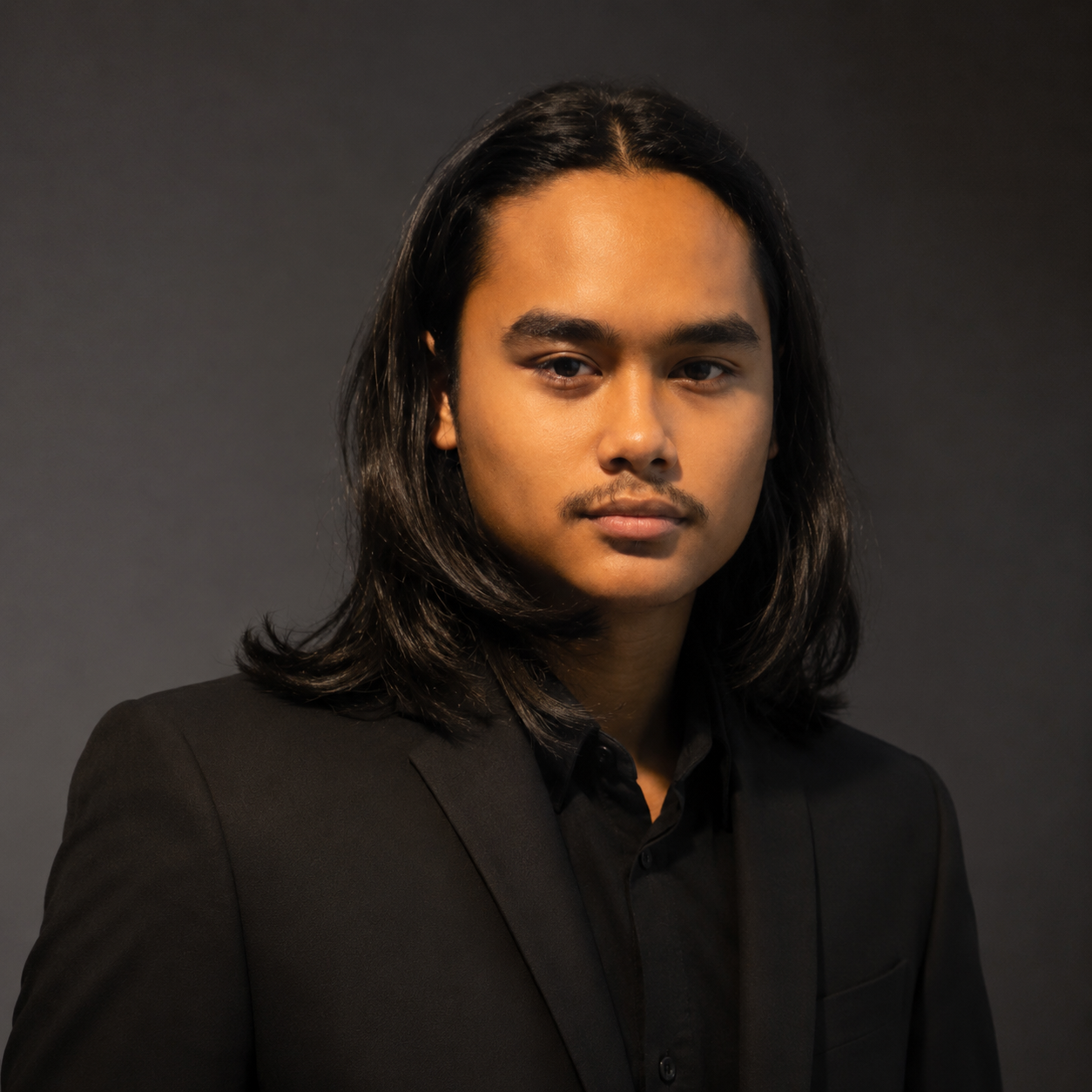 A young man with long black hair, dressed in a black suit, looking directly at the camera against a dark background.