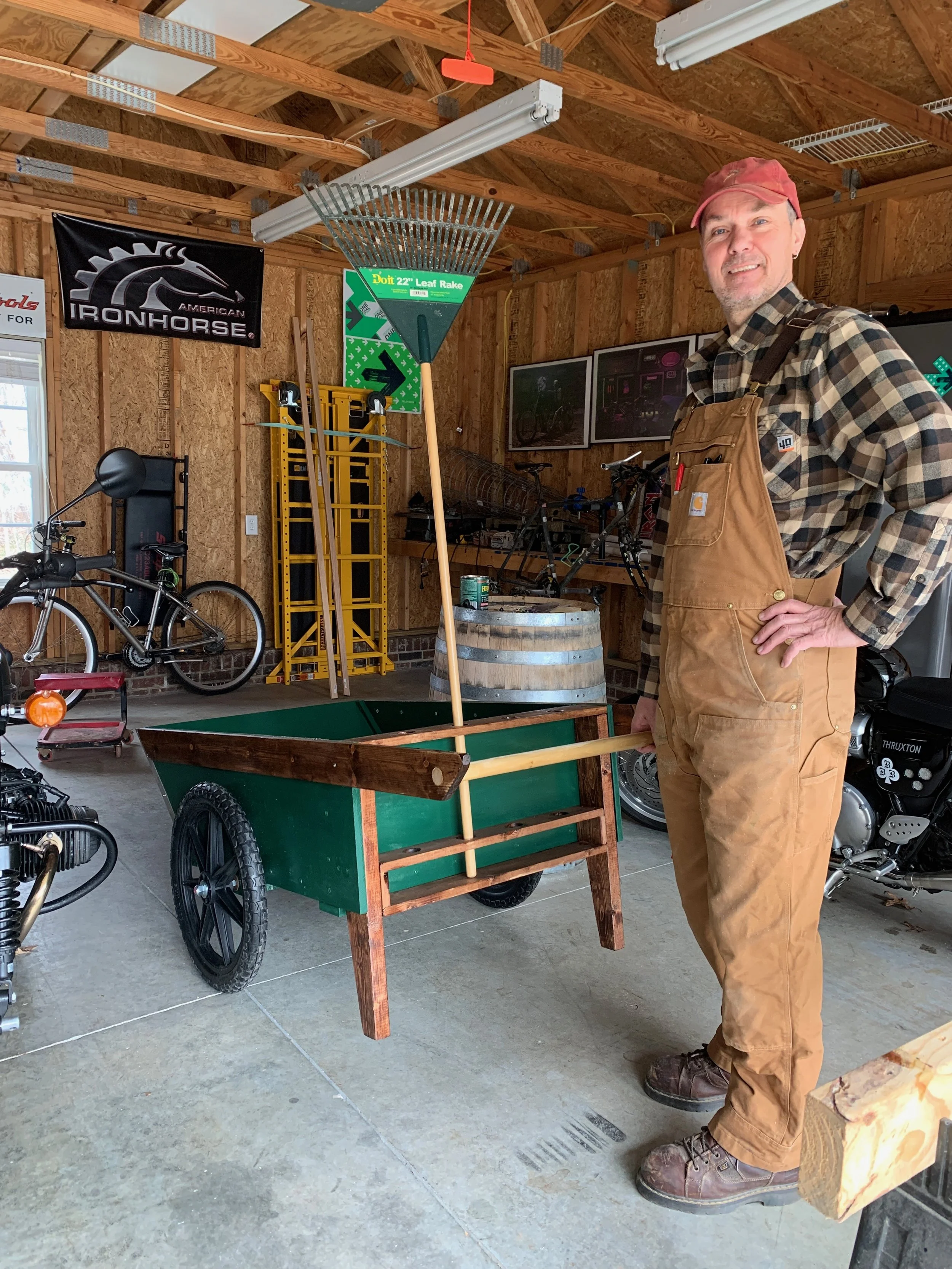 A man wearing a red baseball cap and brown overalls stands in a garage or workshop, smiling and posing for the photo. Behind him, there are bicycles, a black banner with a horse and gear logo, a yellow ladder, a barrel, and various woodworking or bicycle tools and picture frames.