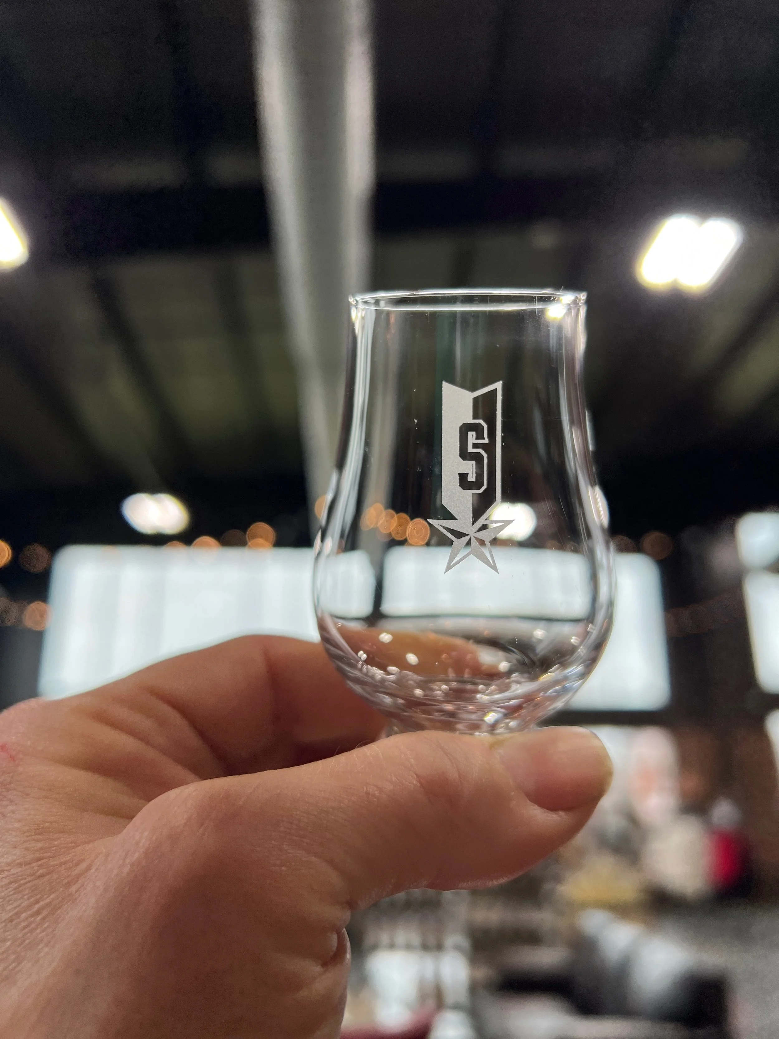 A clear glass held between fingers, with a blurred indoor setting in the background.