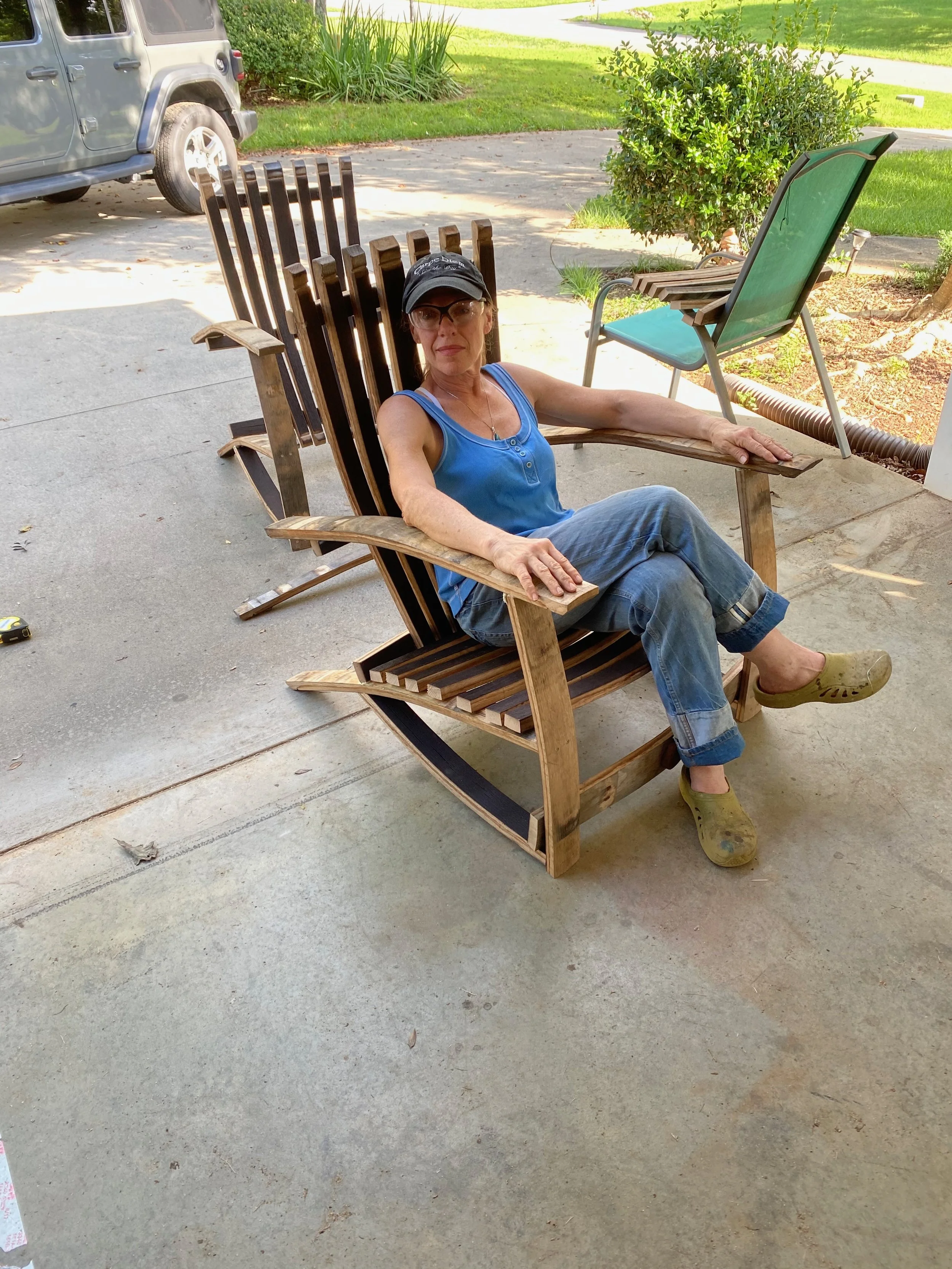 A woman sitting on an unfinished wooden rocking chair outside on a concrete surface, wearing a blue tank top, jeans, green Crocs, glasses, and a black cap.