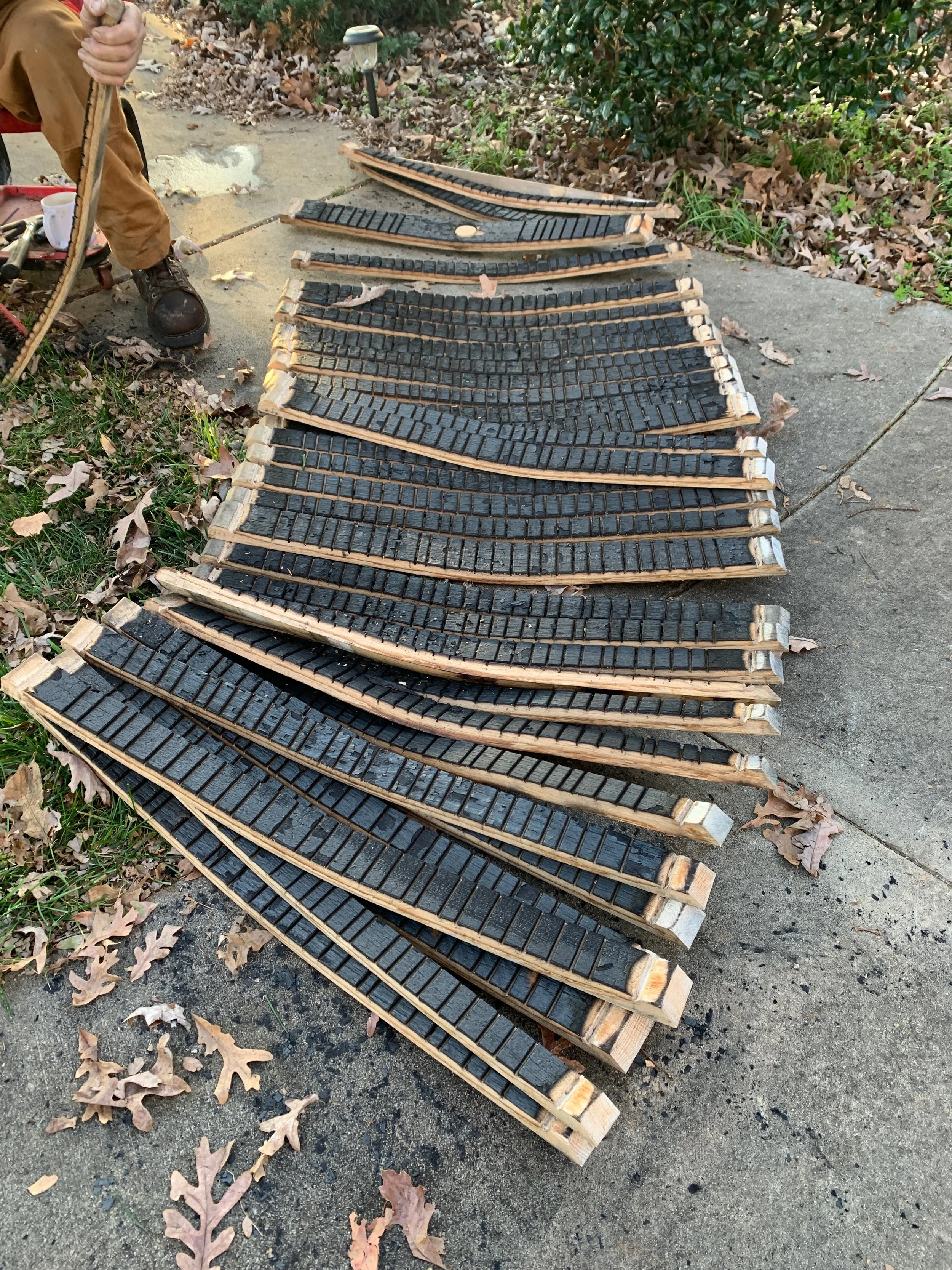 A person working outdoors with weathered wooden frames filled with black material, possibly for a DIY project, on a sidewalk surrounded by fallen leaves and bushes.