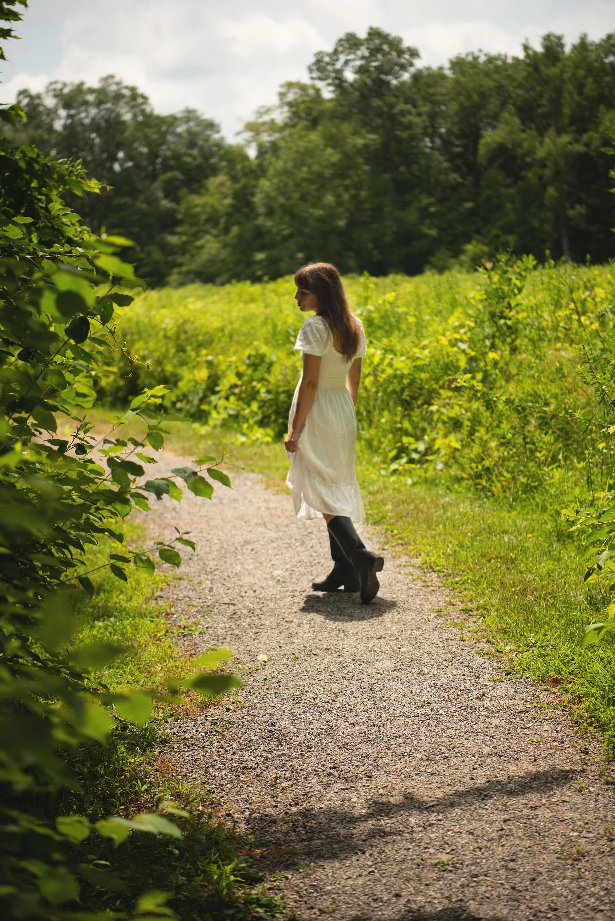 Alexandra Poole LCSW in a white dress and black boots walking on a gravel path surrounded by green bushes and trees under cloudy sky.