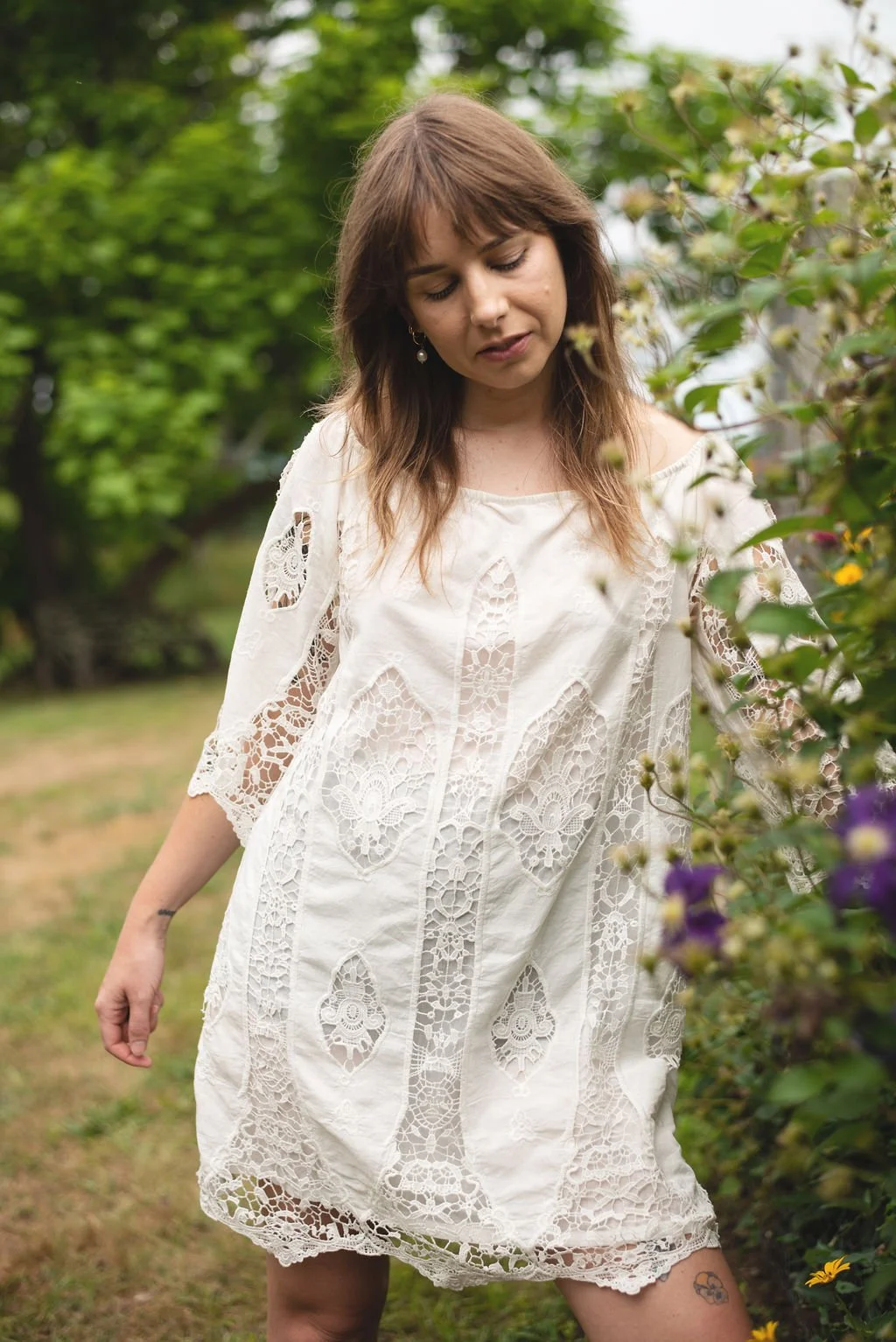 A woman in a white lace dress standing outdoors among greenery and flowers, looking down.