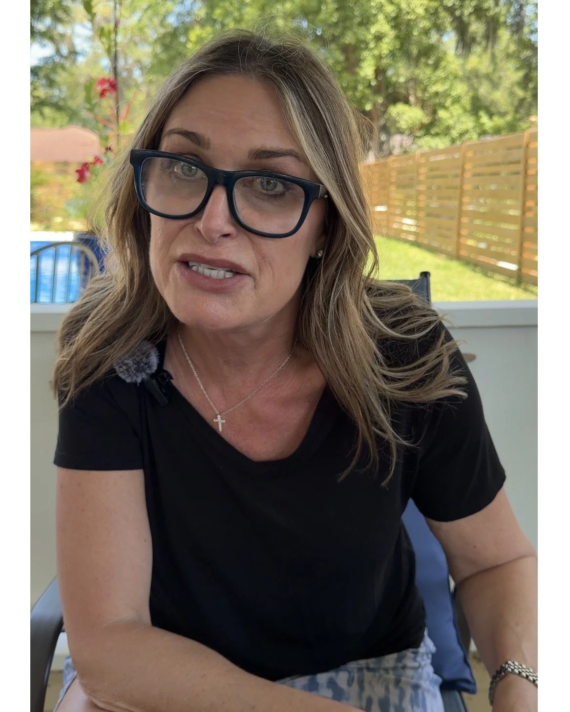 A middle-aged woman with light brown hair and glasses sitting outdoors on a porch with green trees and a wooden fence in the background.