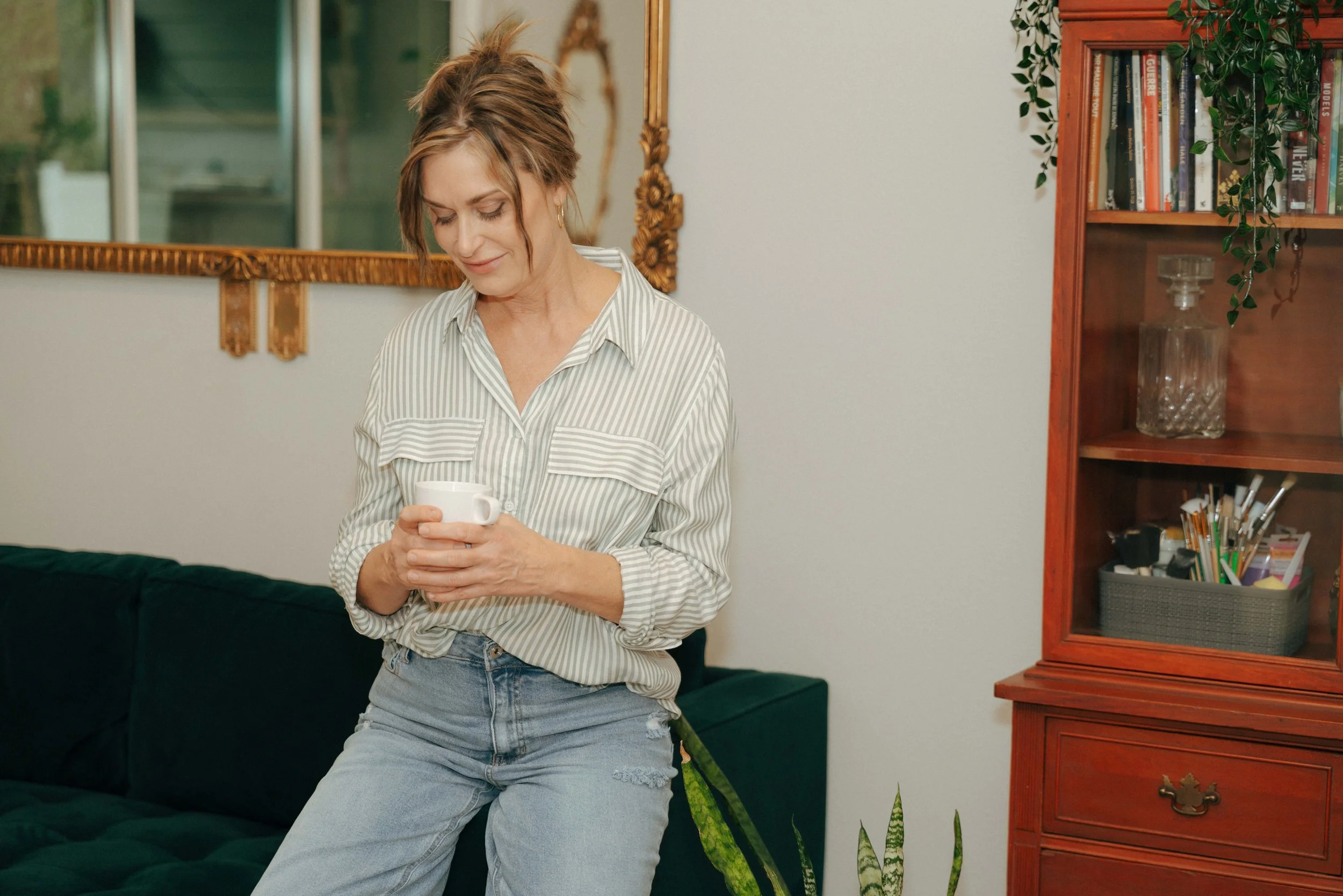 A woman with short, tousled hair wearing a striped shirt and ripped jeans, sitting on a green couch, holding a white mug, in a room with a wooden bookshelf, a mirror, and some green plants.