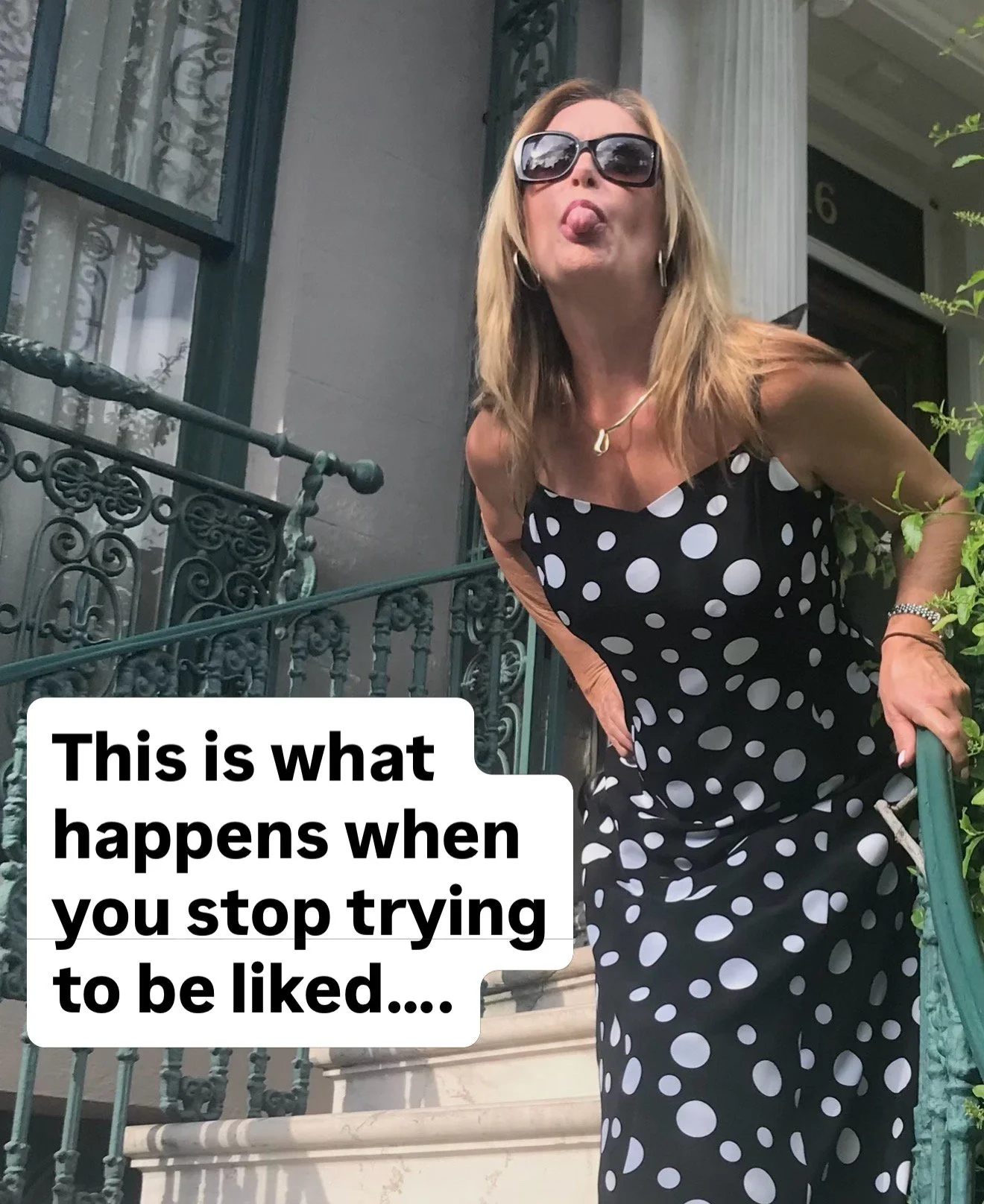 A woman wearing sunglasses and a black dress with white polka dots standing on the steps of a house with ornate black railings, sticking out her tongue, with a humorous caption that reads 'This is what happens when you stop trying to be liked....'