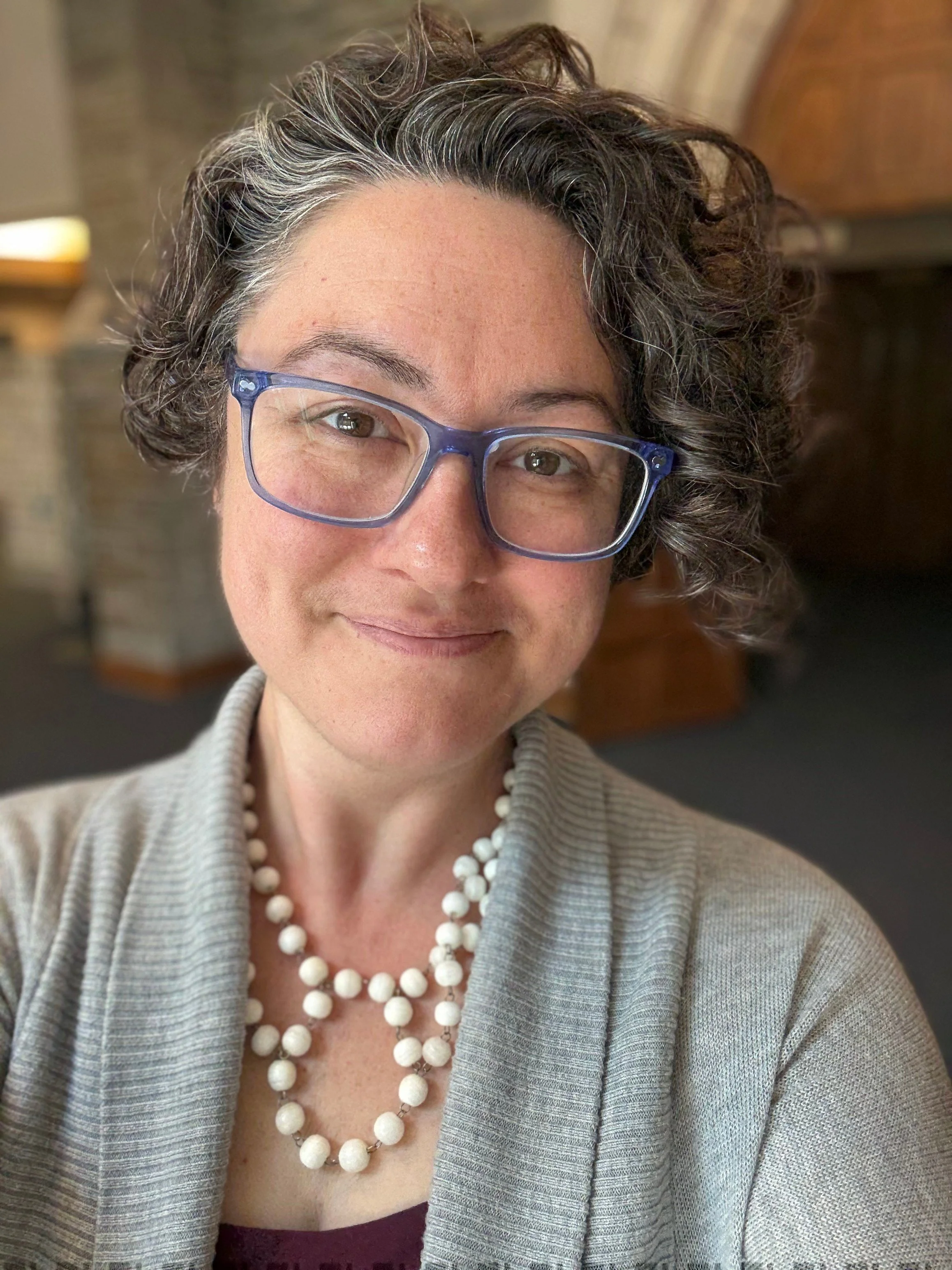 Close-up of a smiling woman with curly gray hair, wearing blue glasses, a gray cardigan, and a pearl necklace.