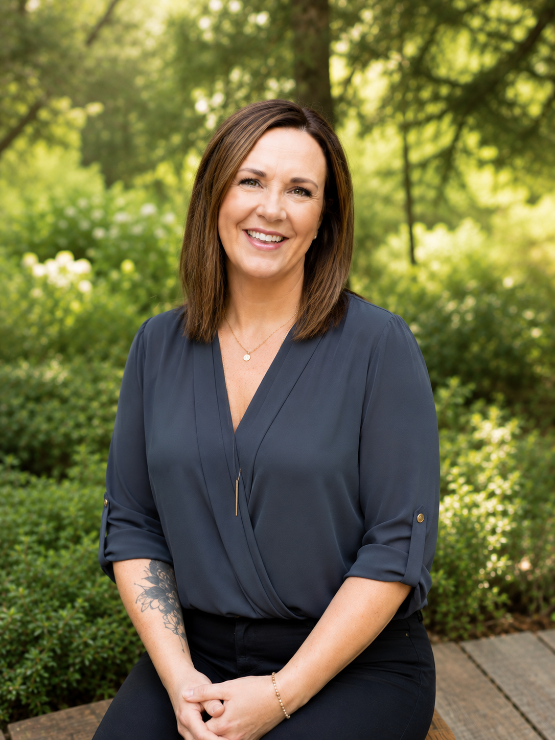 A woman with shoulder-length brown hair smiling outdoors in front of a green, leafy background.