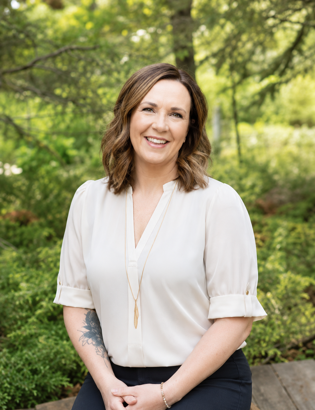 A woman with shoulder-length wavy brown hair, wearing a white blouse with rolled-up sleeves, smiling, sitting outdoors in a lush green park.