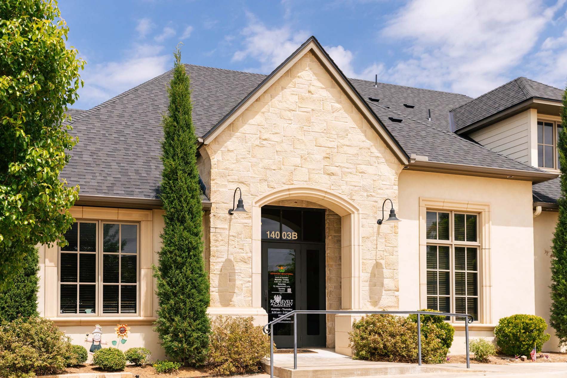 Front view of a modern buildingwith a stone facade, large windows, a dark front door with a sign, and a landscaped garden with shrubs and small American flags. The house has a multi-gabled roof with gray shingles and a clear sky in the background.