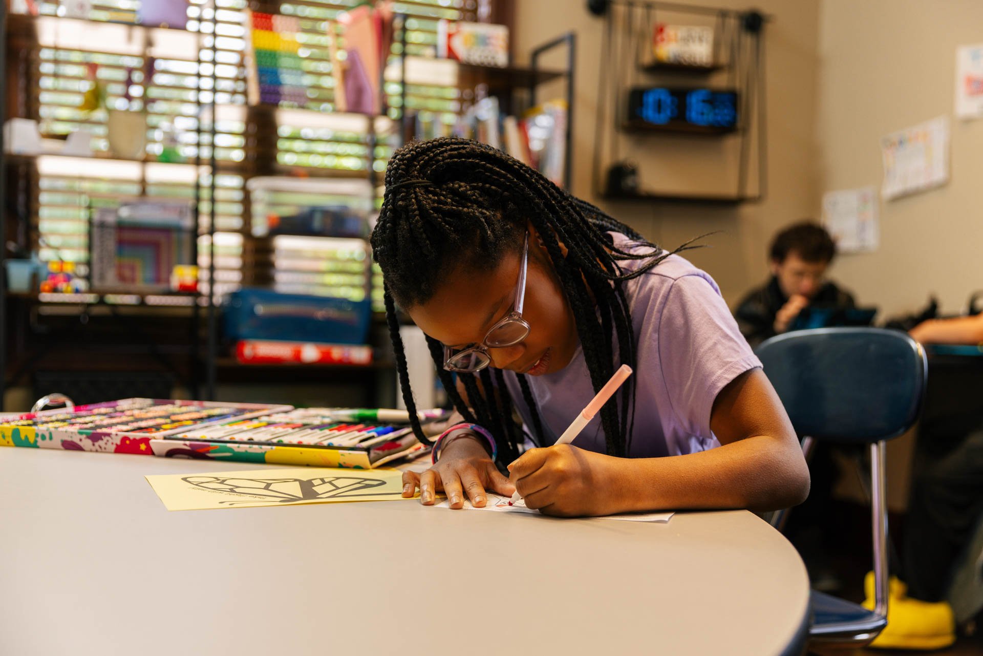 Young girl with braided hair and glasses coloring a picture at a table in a classroom with shelves and a clock in the background.