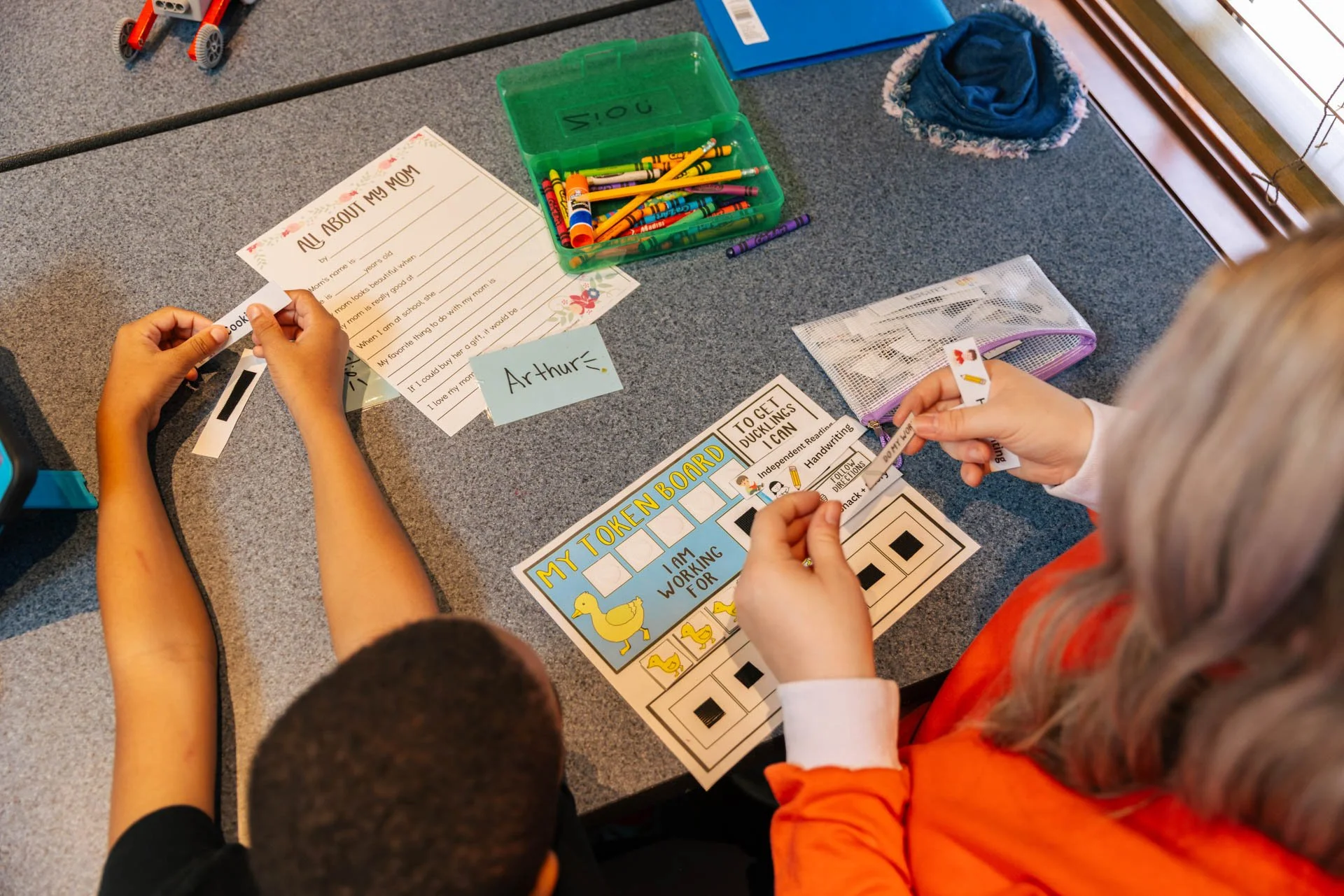 Children working on a classroom activity involving writing and matching. One child is holding a strip of paper with a picture and another child is placing it on a worksheet. The worksheet has a title "My Token Board" with images of ducks and writing prompts.